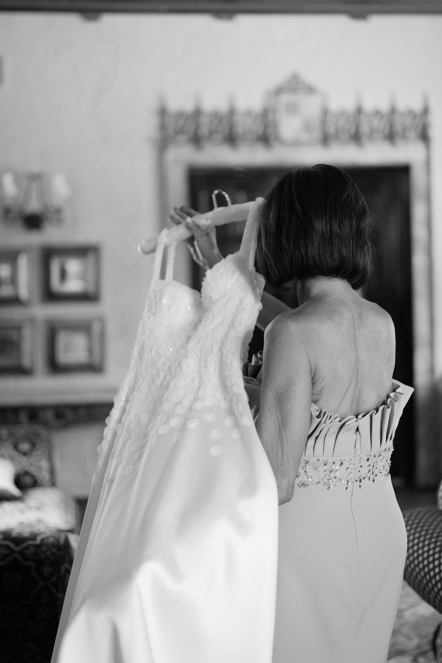 Black and white photo of a bride holding her lace wedding dress on a hanger in an elegant room.