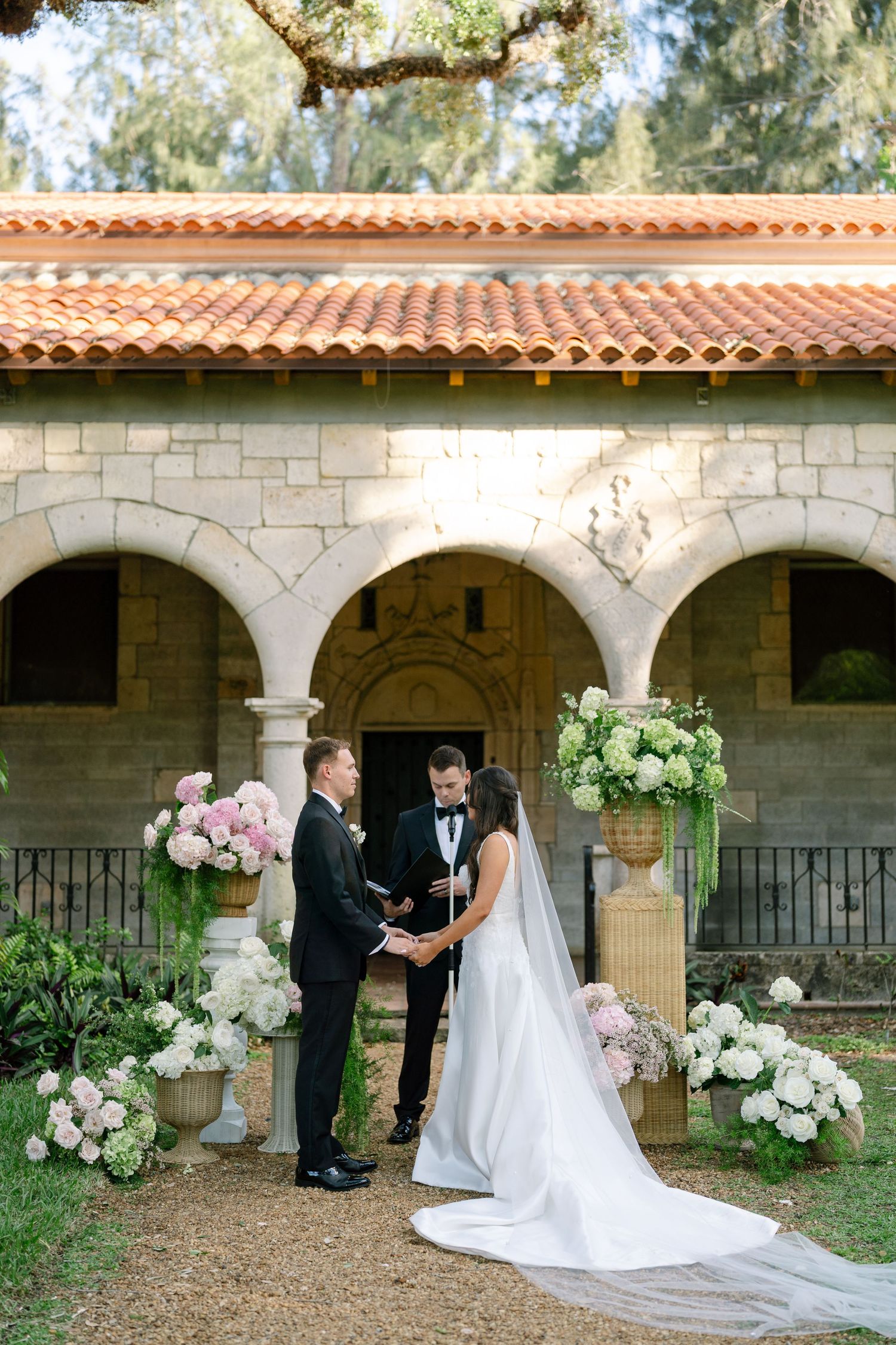 Bride and groom exchange vows before stone chapel with pink and white floral arrangements at outdoor wedding.