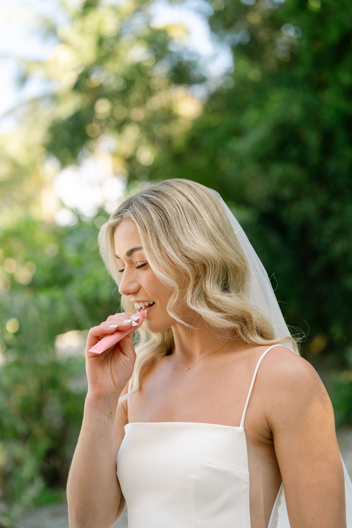 A bride in a simple white dress smiling while touching her face outdoors.