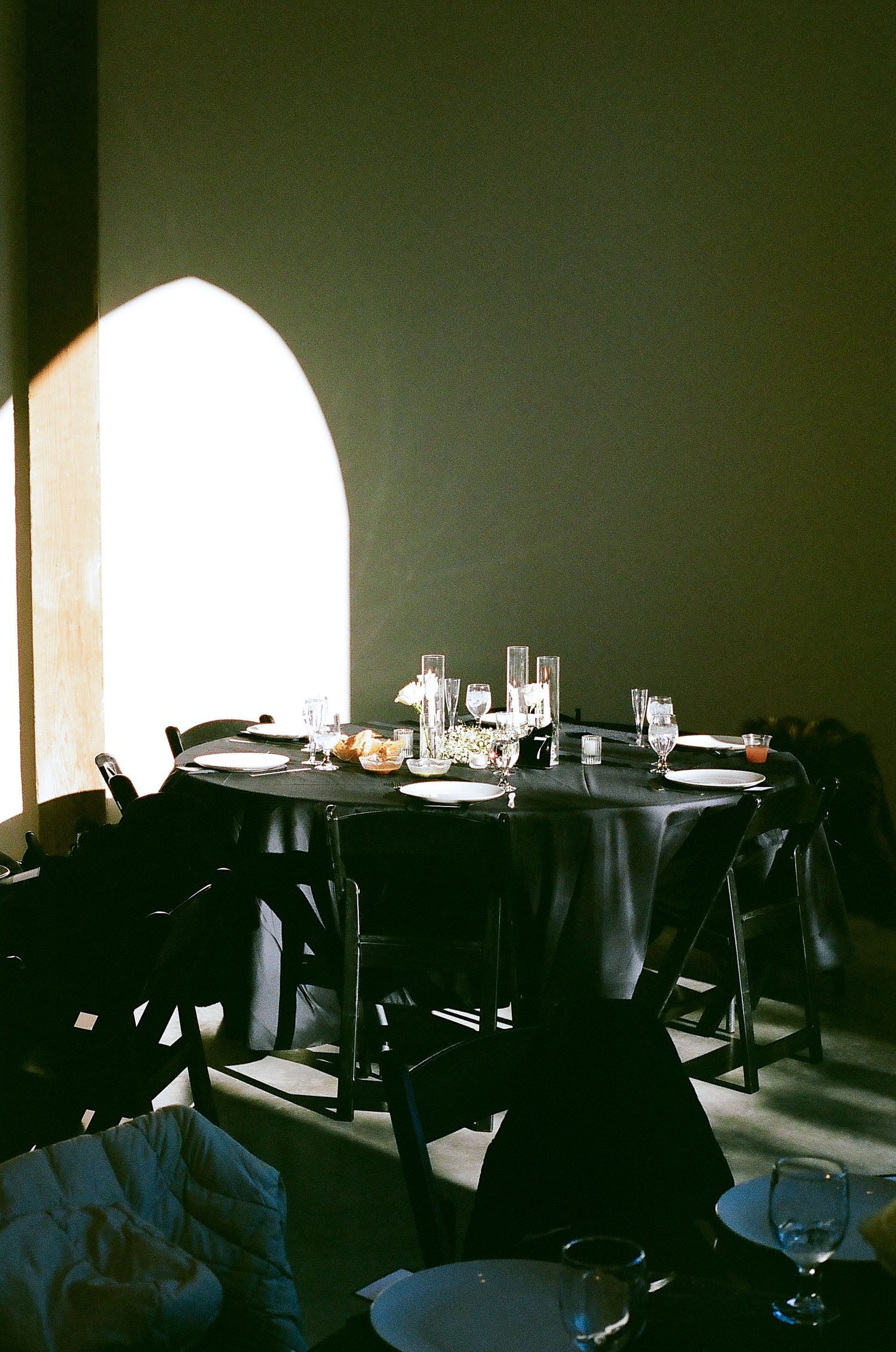 Dark restaurant interior with tables set for dining and arched windows casting dramatic natural light into the space.