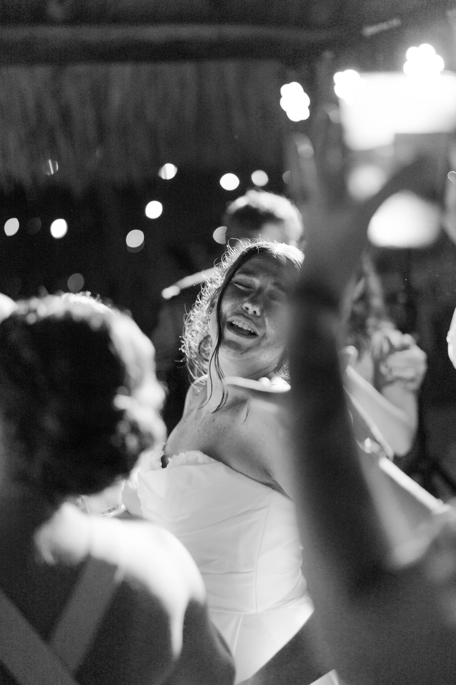 Black and white photo of a bride dancing joyfully at her wedding reception under twinkling lights.