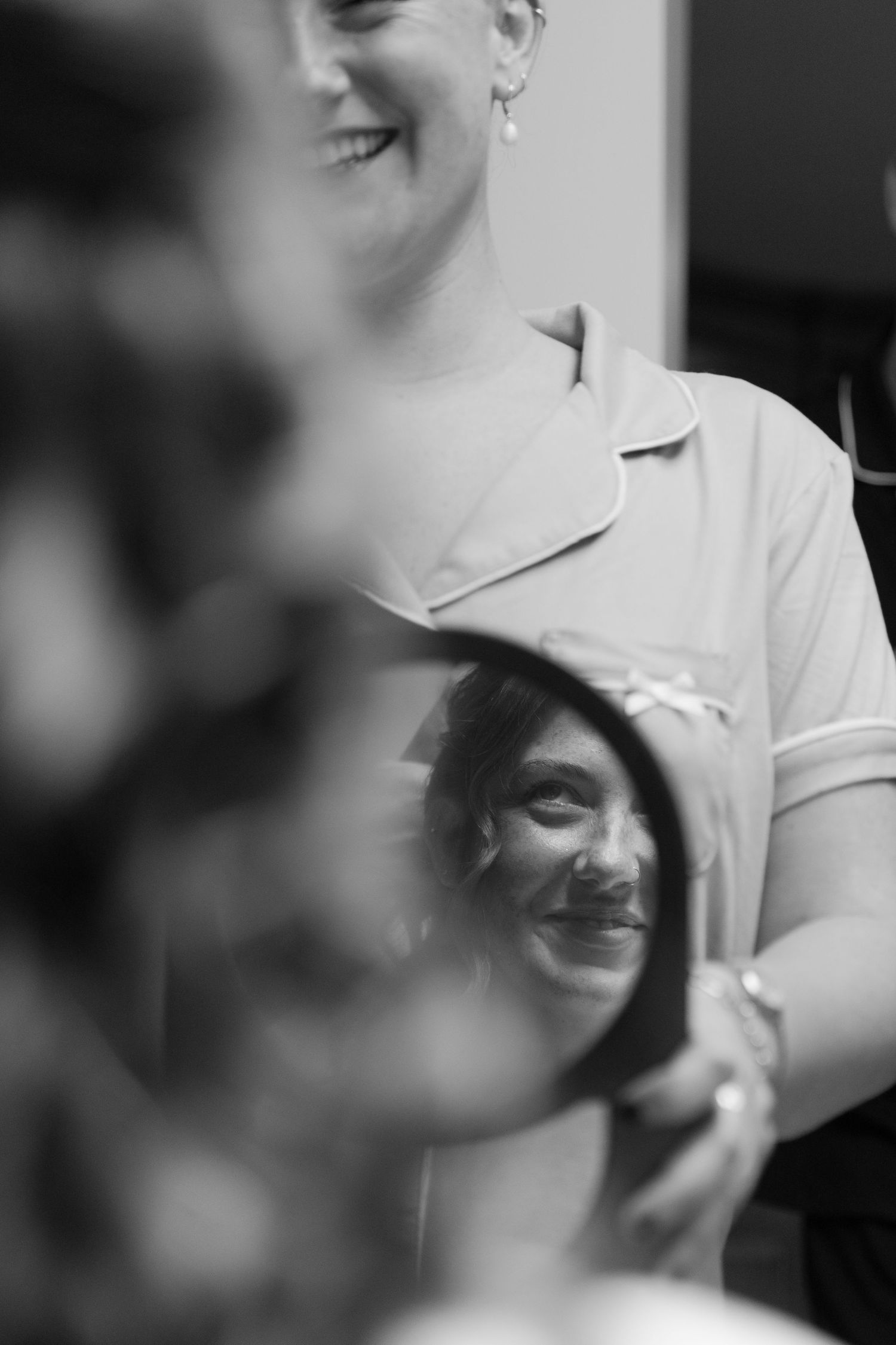 A black and white photograph shows a woman smiling and looking at her reflection in a mirror.