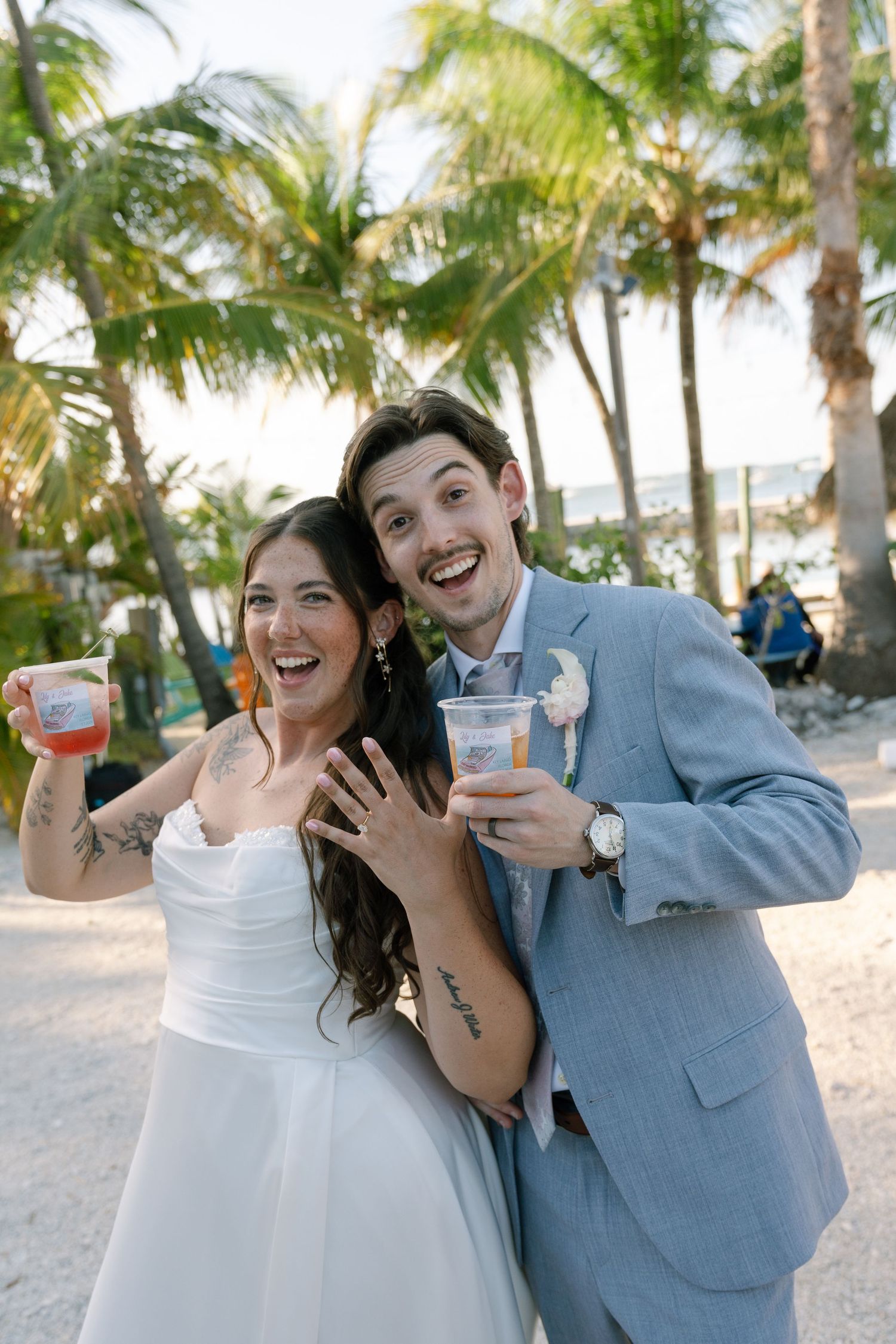 Joyful newlyweds celebrate on a tropical beach, bride showing ring, both holding drinks, palm trees in background.