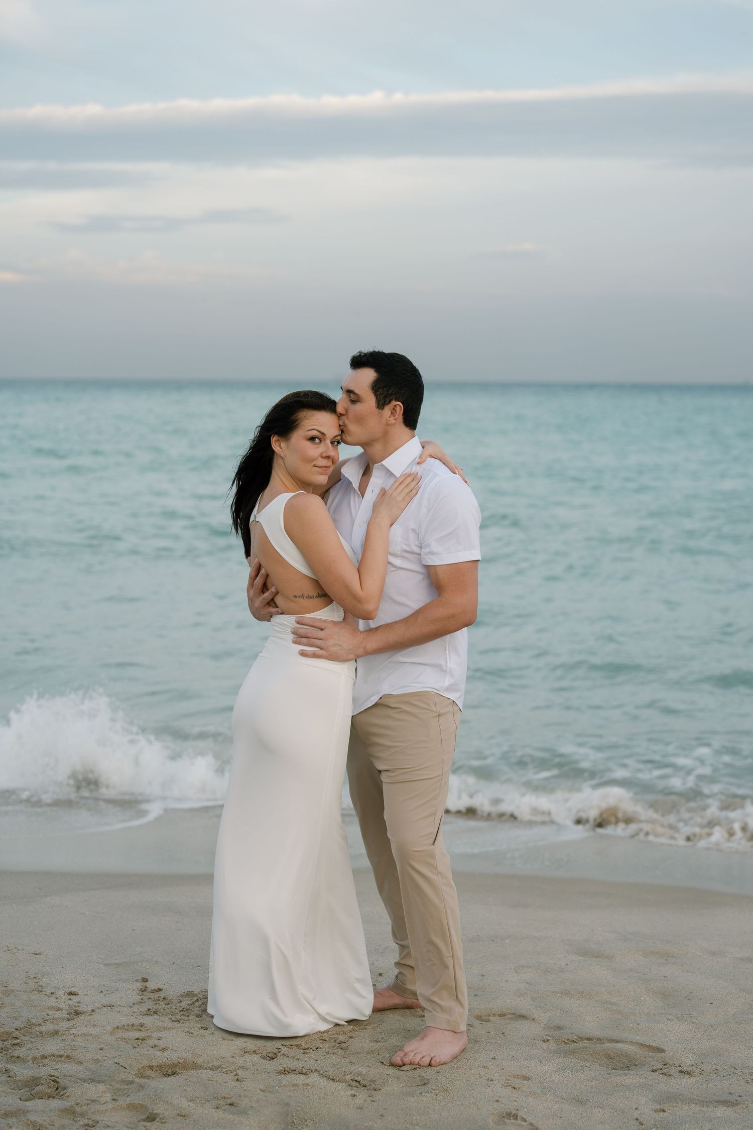 Couple in white and khaki sharing intimate moments on a beach with turquoise waters.