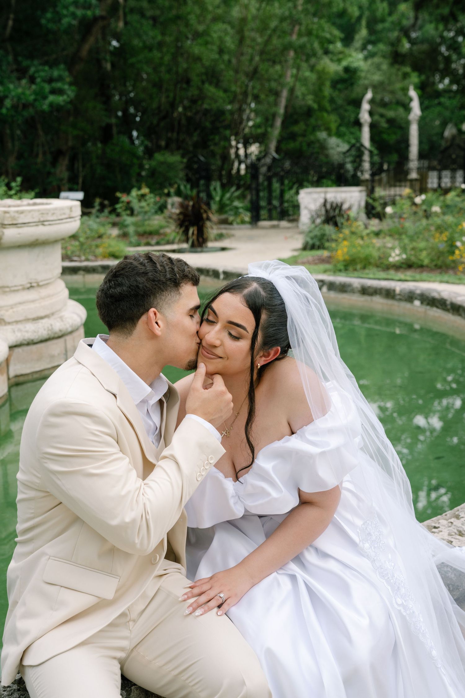 A couple in formal wedding attire share intimate moments in a romantic garden setting with stone columns and greenery.