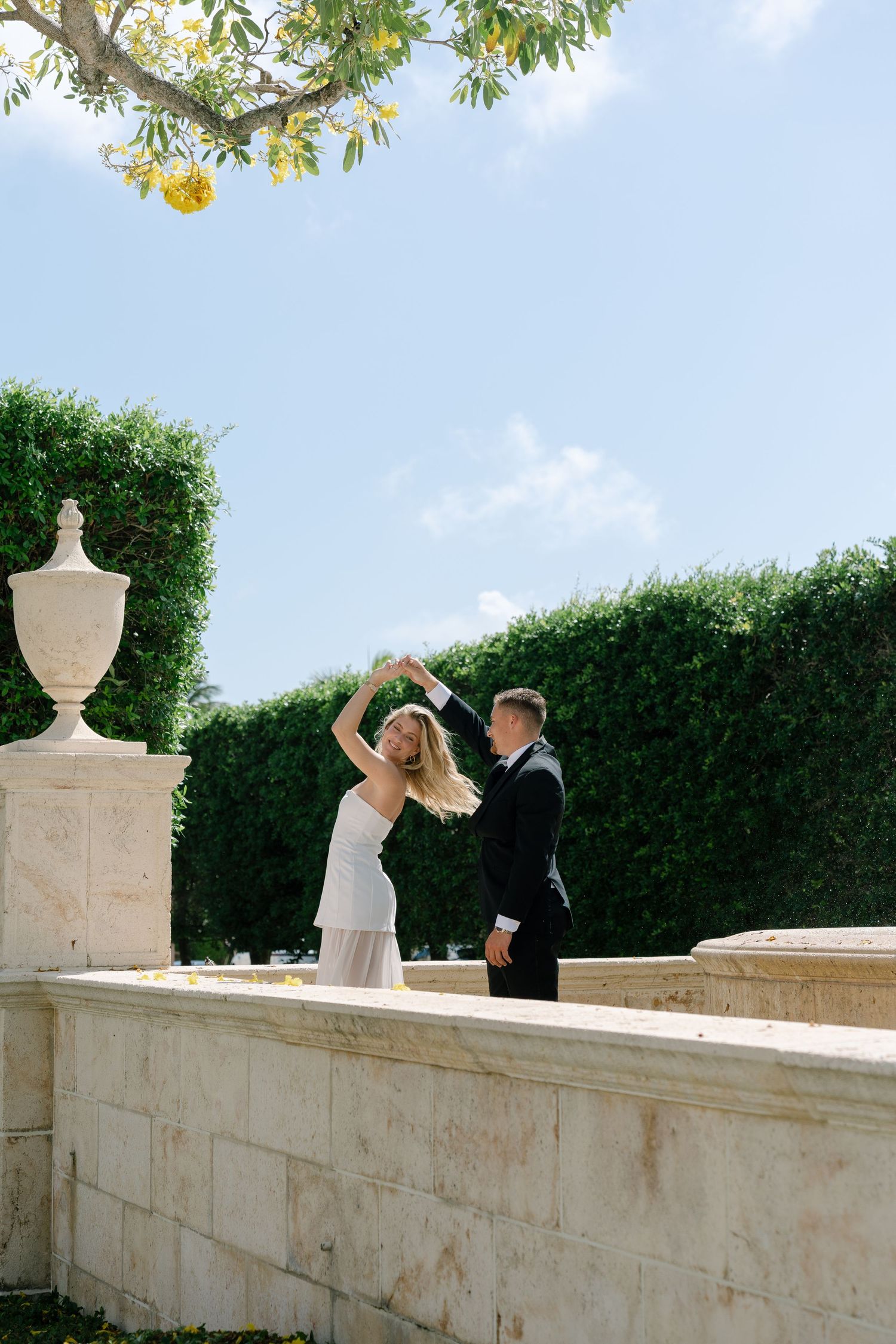 Bride and groom dancing outdoors near elegant stone wall with lush green hedges and decorative urns under blue sky.