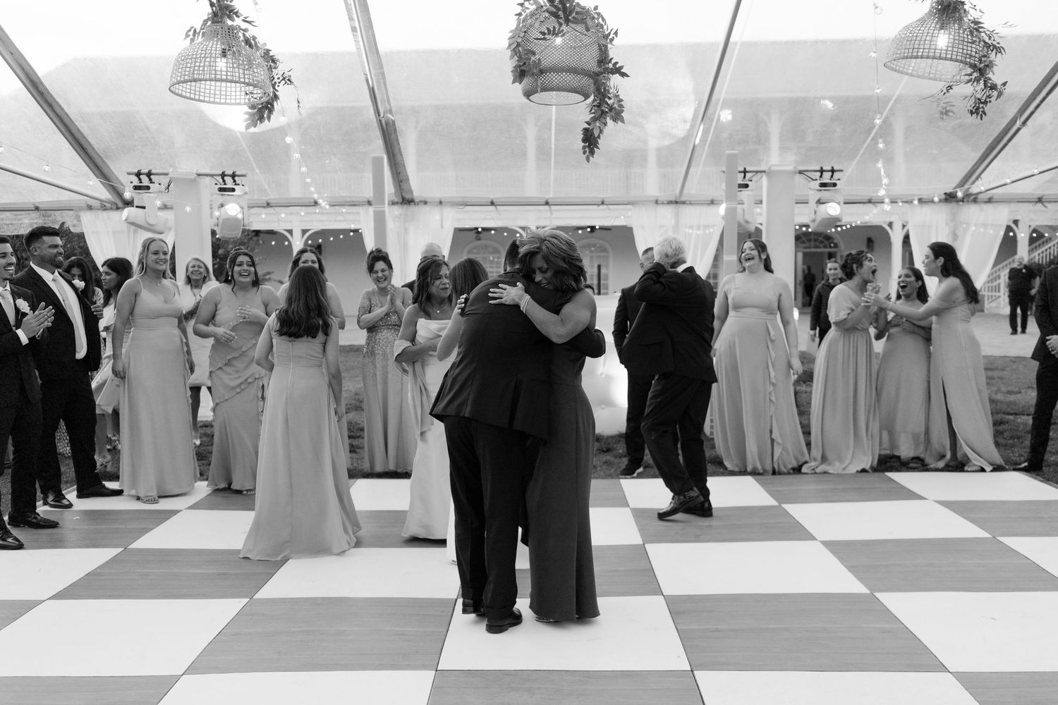 Couple sharing emotional hug on checkered dance floor surrounded by bridesmaids in tent reception.