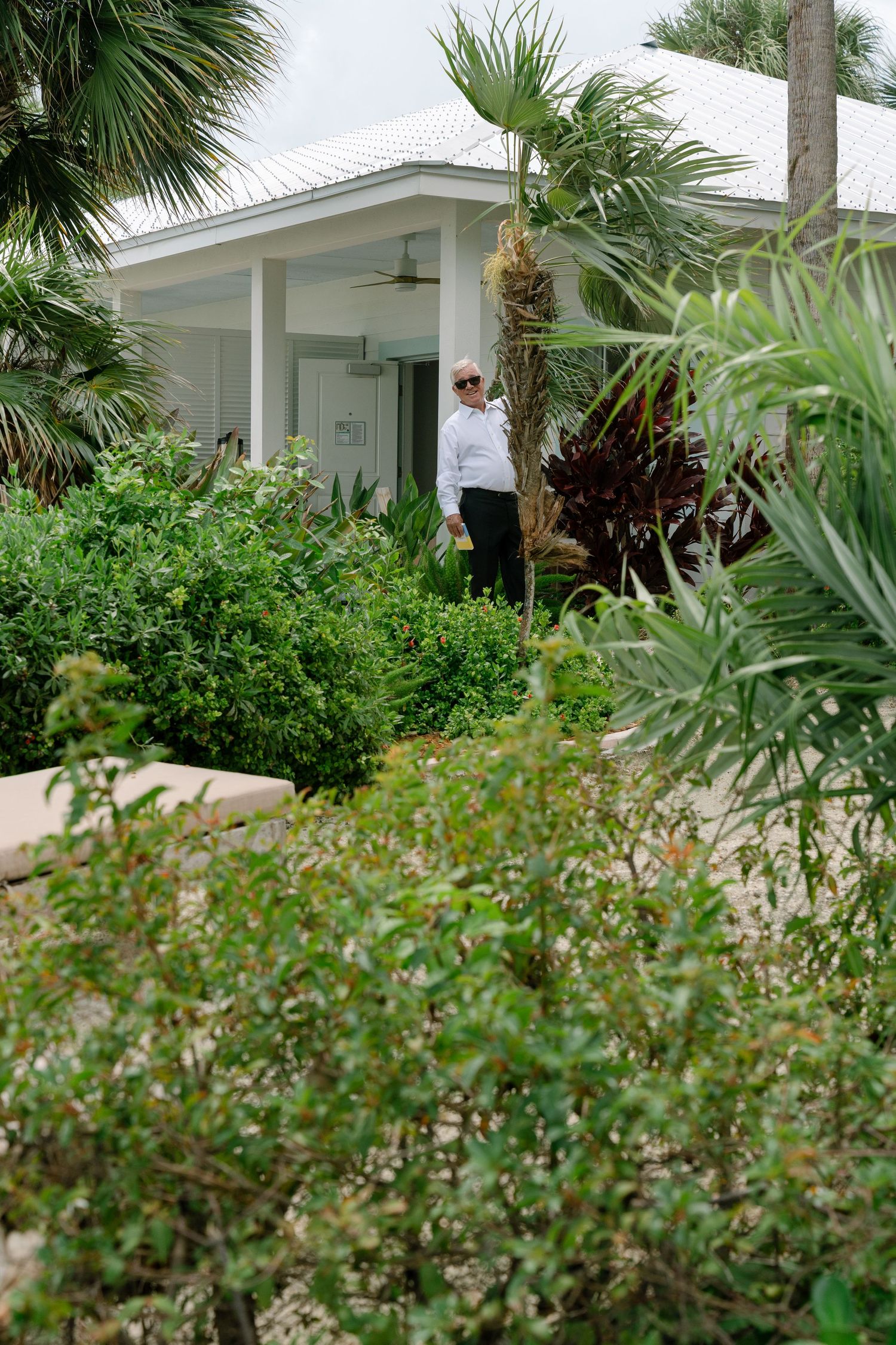 A person in a white shirt stands among lush tropical greenery outside a white cottage with palm trees.