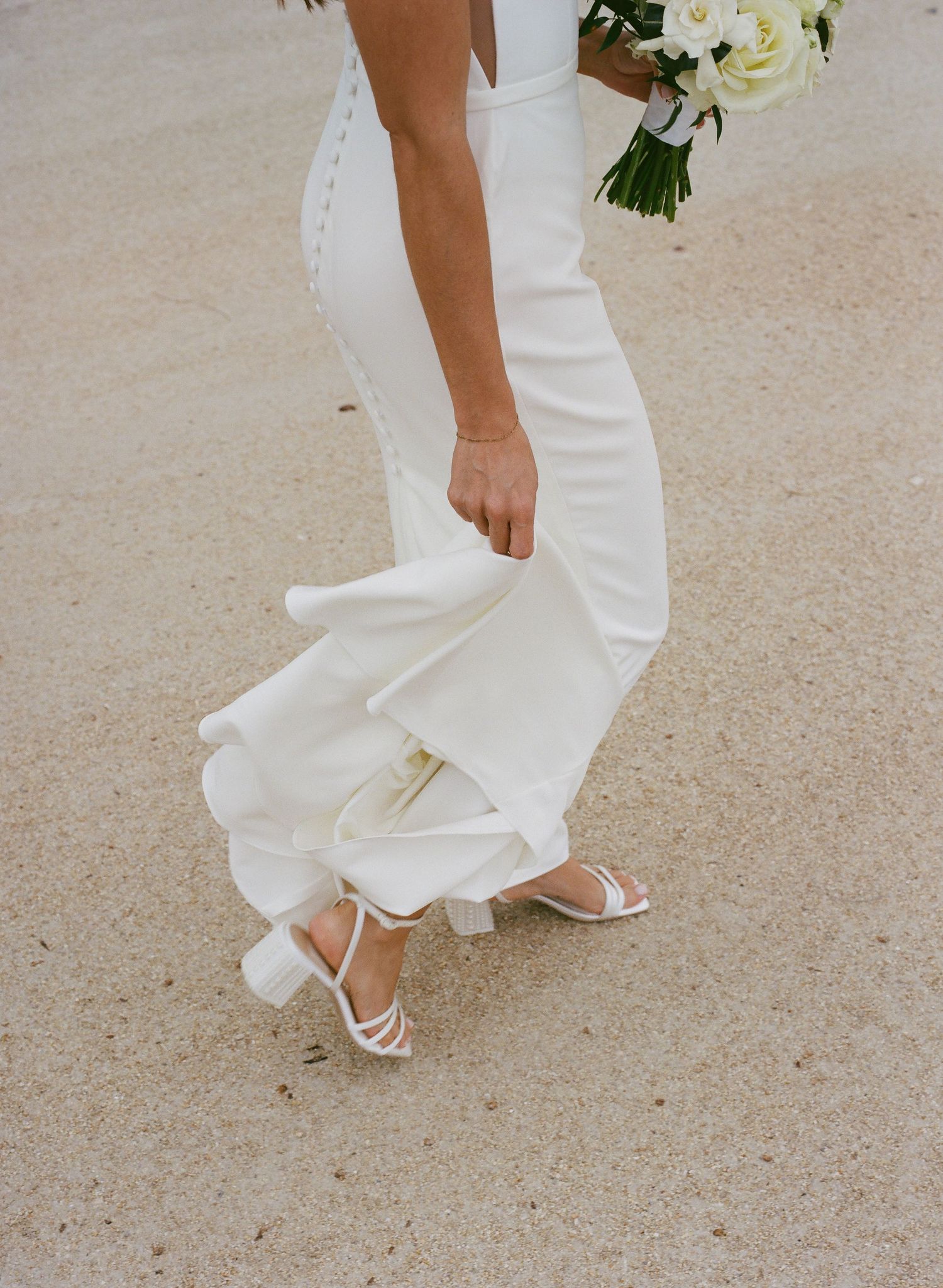 Bride in white wedding gown lifting dress hem, wearing strappy white heels, holding bouquet on sandy ground.