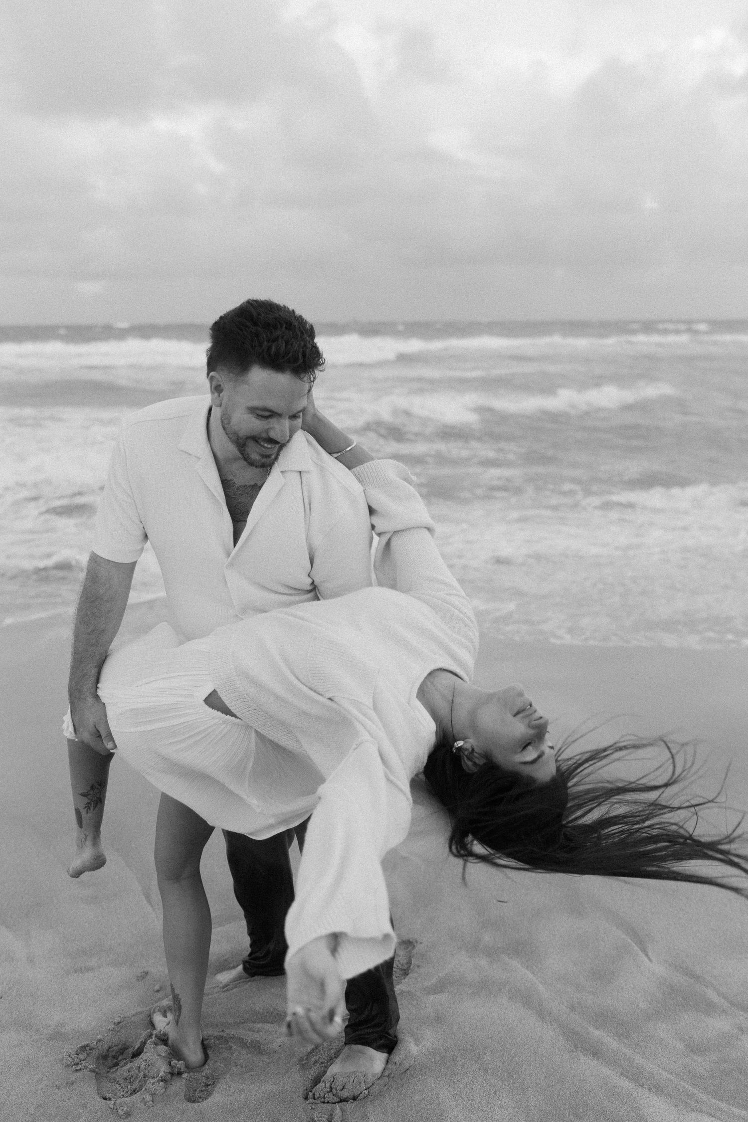 A couple in white clothing playfully poses on a beach with waves in the background.