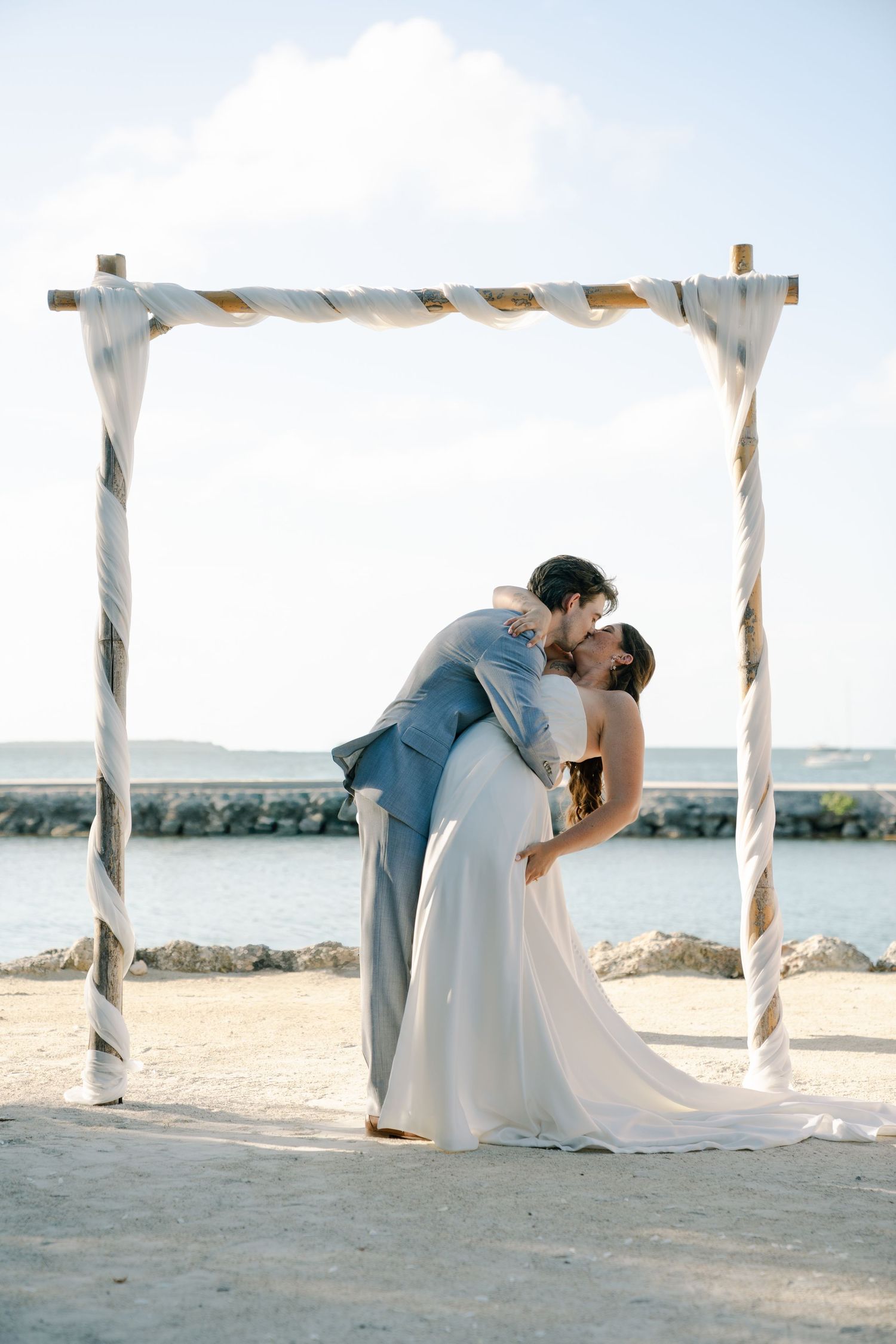 The image shows a bride and groom embracing under a decorated arch on a beach.