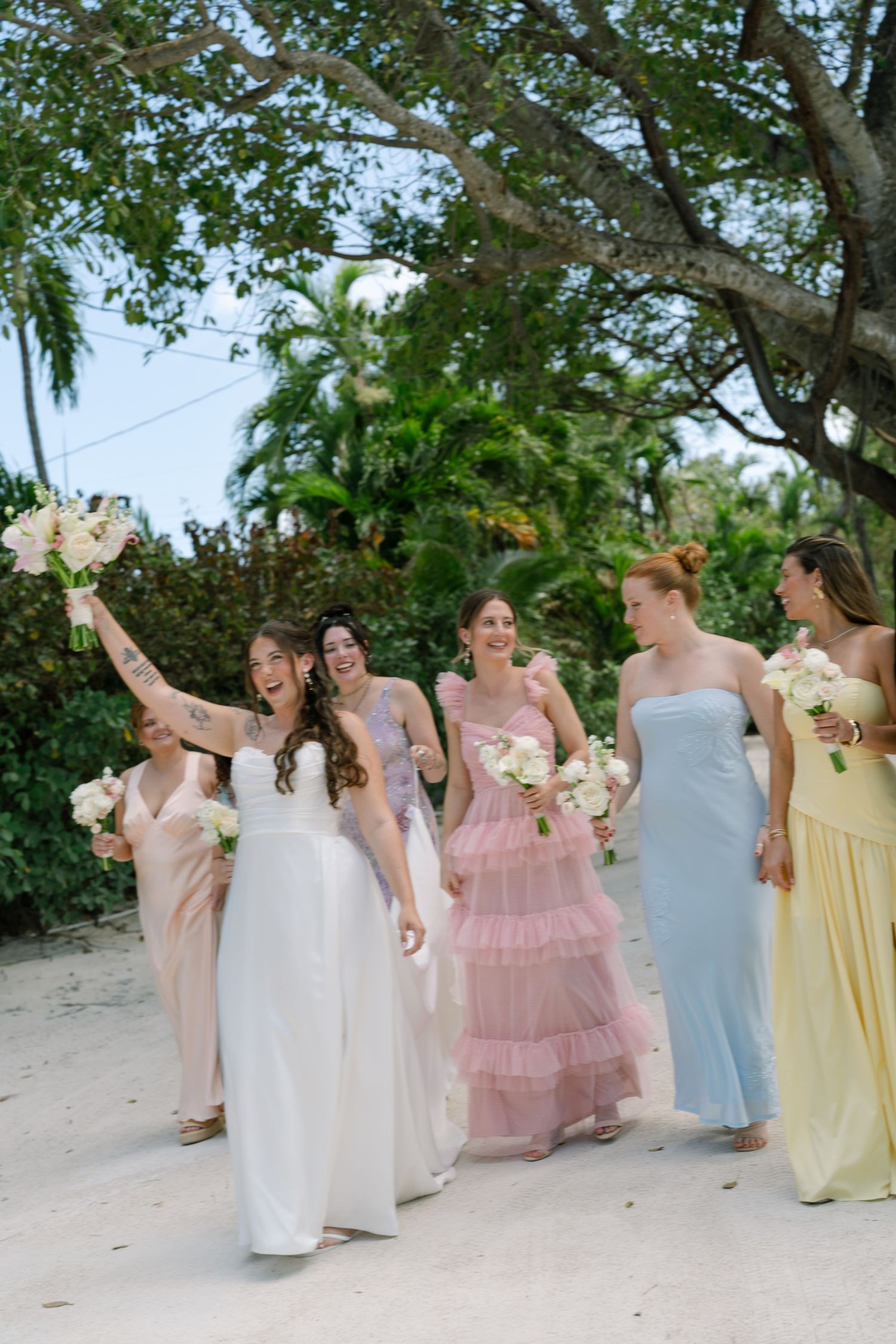 A group of bridesmaids in pastel dresses walk together on a beach.