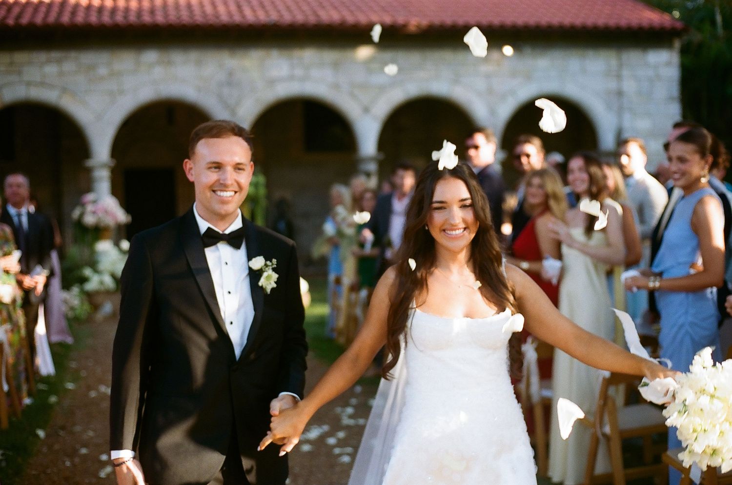 Happy newlyweds walk down the aisle as guests toss white flower petals at an elegant outdoor wedding ceremony.