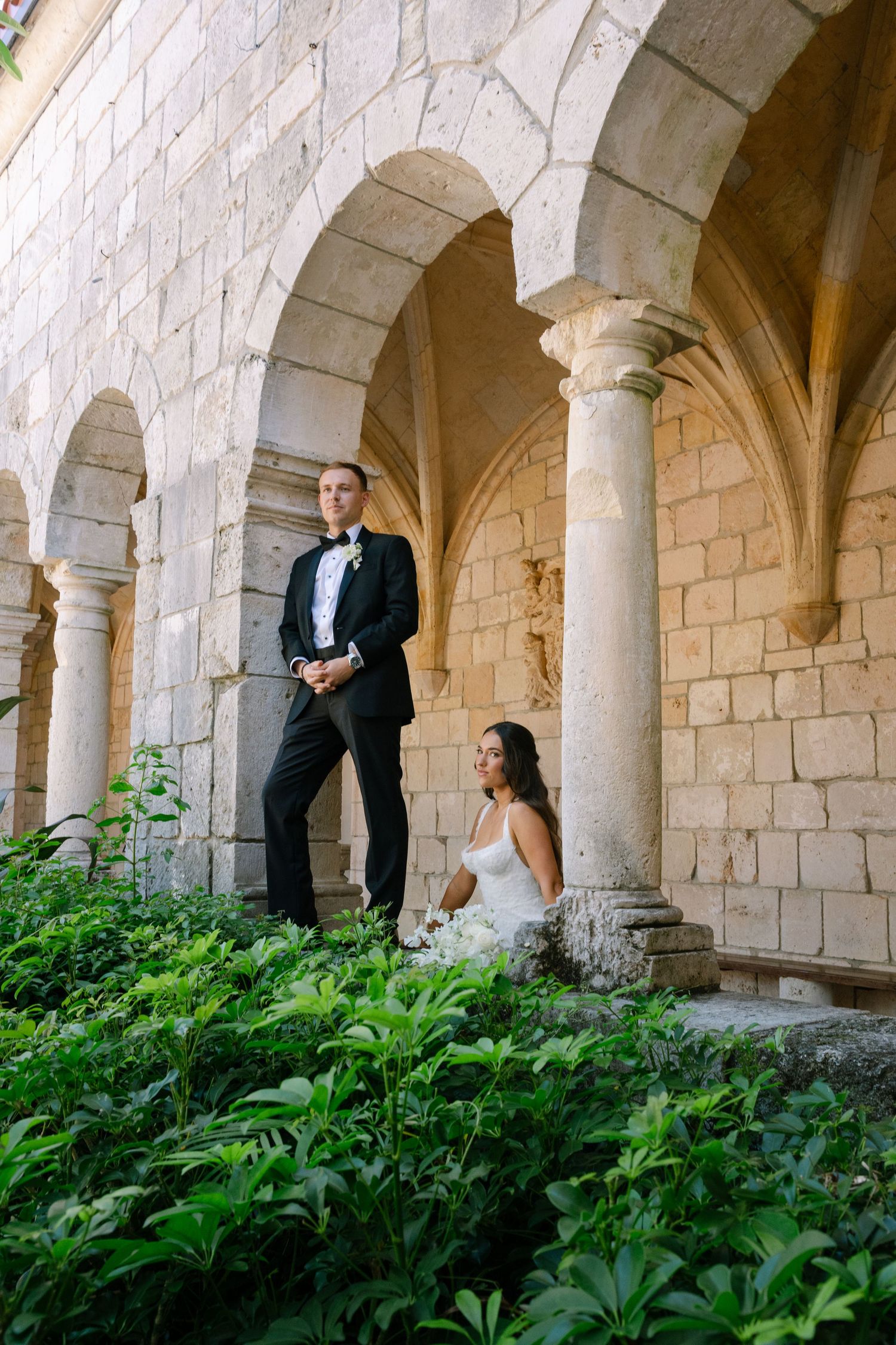 Bride and groom posing among stone arches and lush greenery in an elegant historic venue.