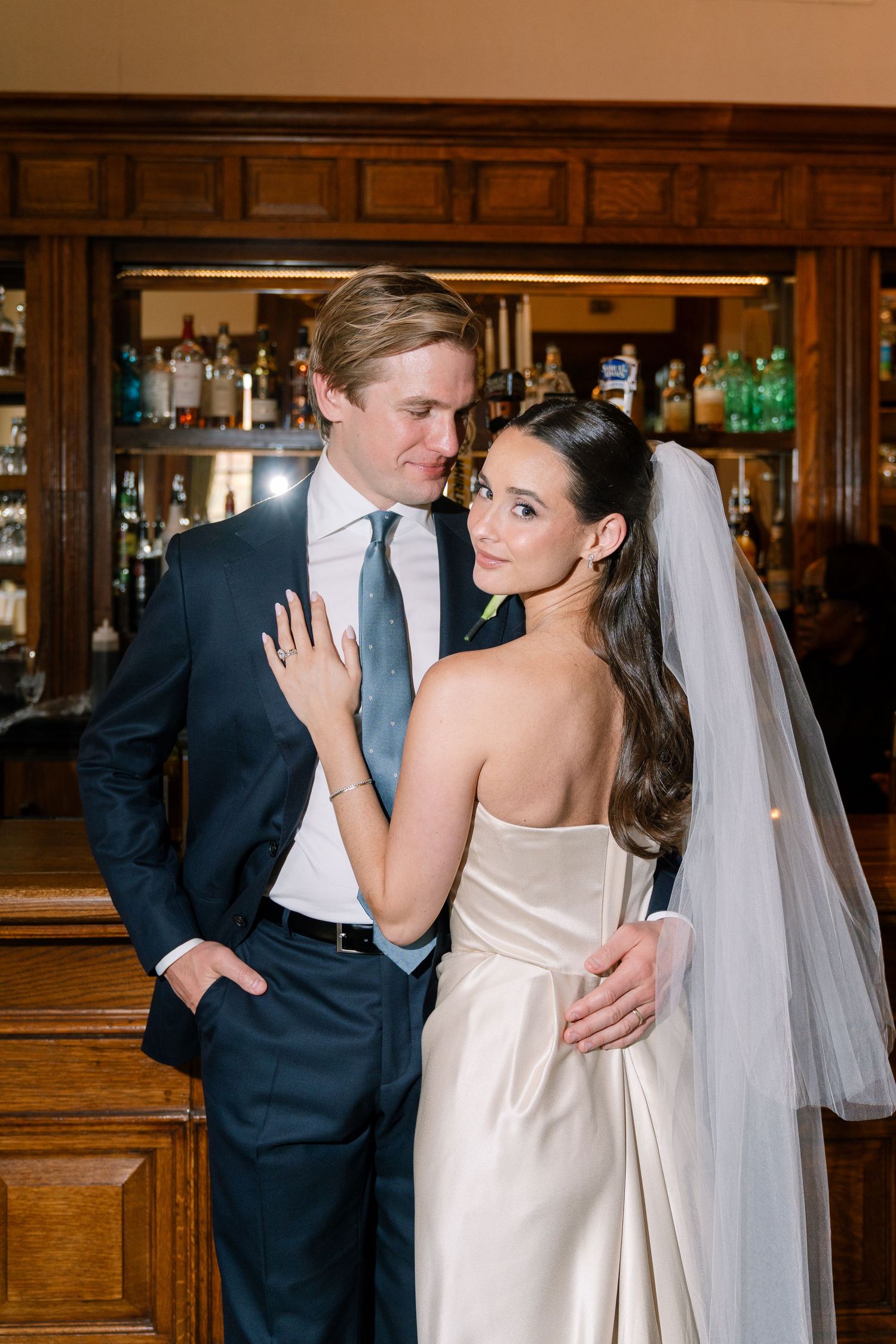 Bride in strapless gown and veil embraces groom in navy suit with blue tie, posing elegantly in front of a wooden bar.