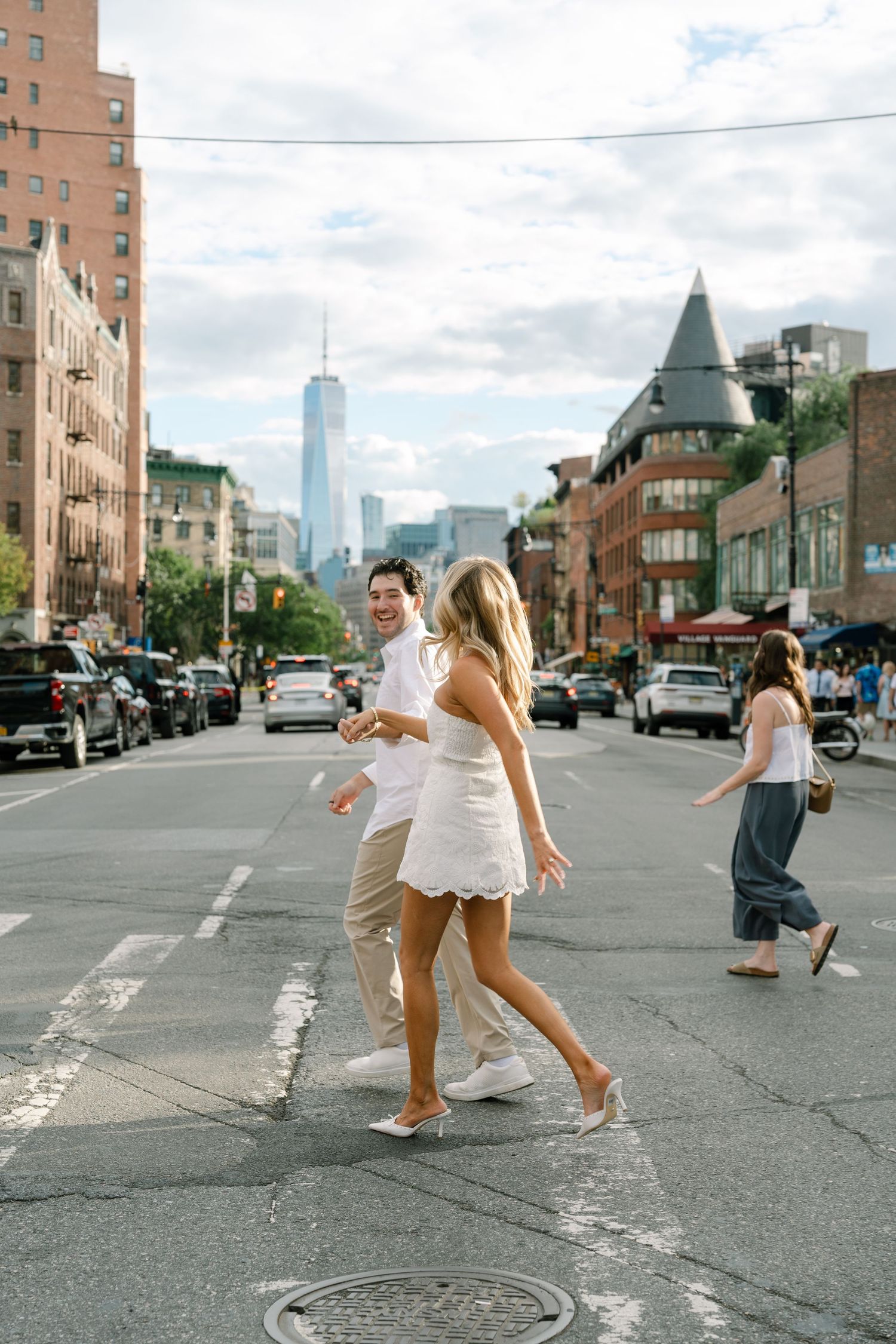 Couple in white outfits joyfully crossing a NYC street with One World Trade Center visible in the background.