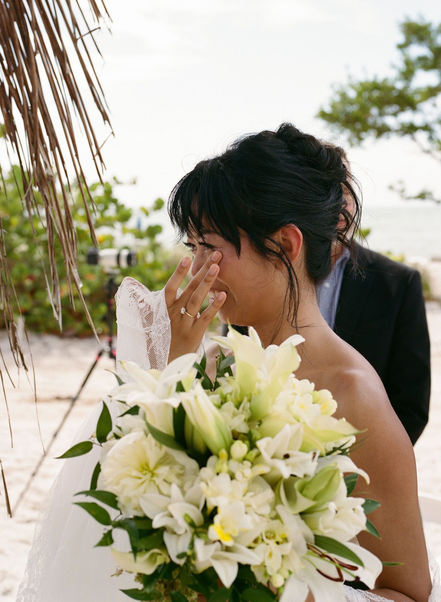 Emotional bride crying while holding white lily bouquet during outdoor beach wedding ceremony.