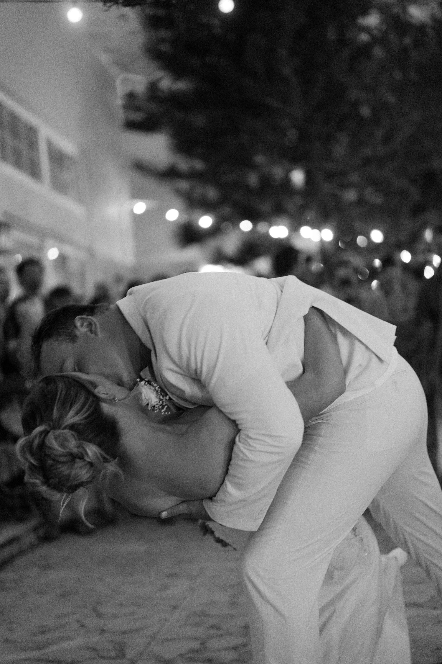 Black and white photo of couple sharing romantic dip kiss at outdoor wedding reception with string lights.