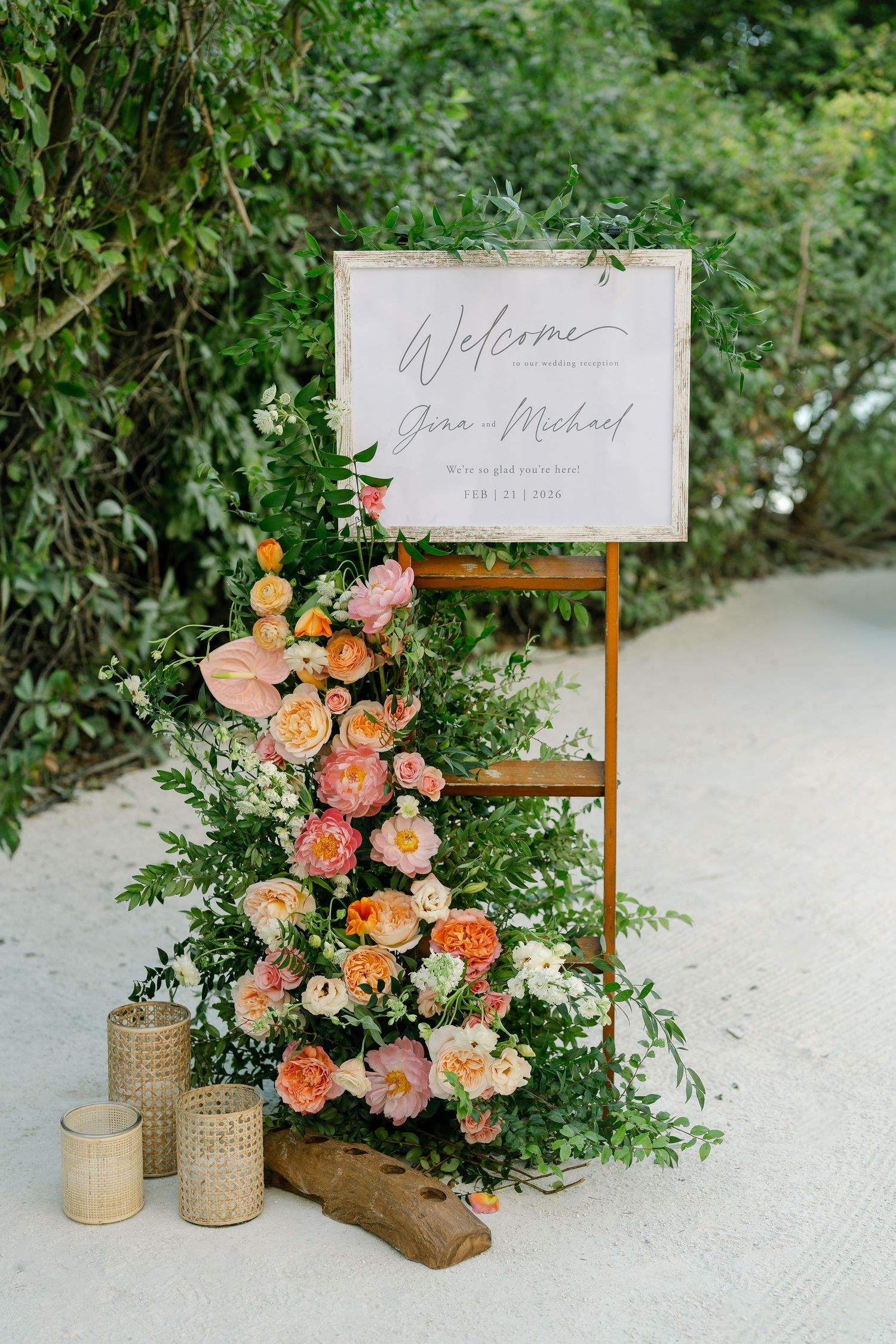 Elegant wedding welcome sign display with coral and peach floral arrangements and woven basket decor on white sand.