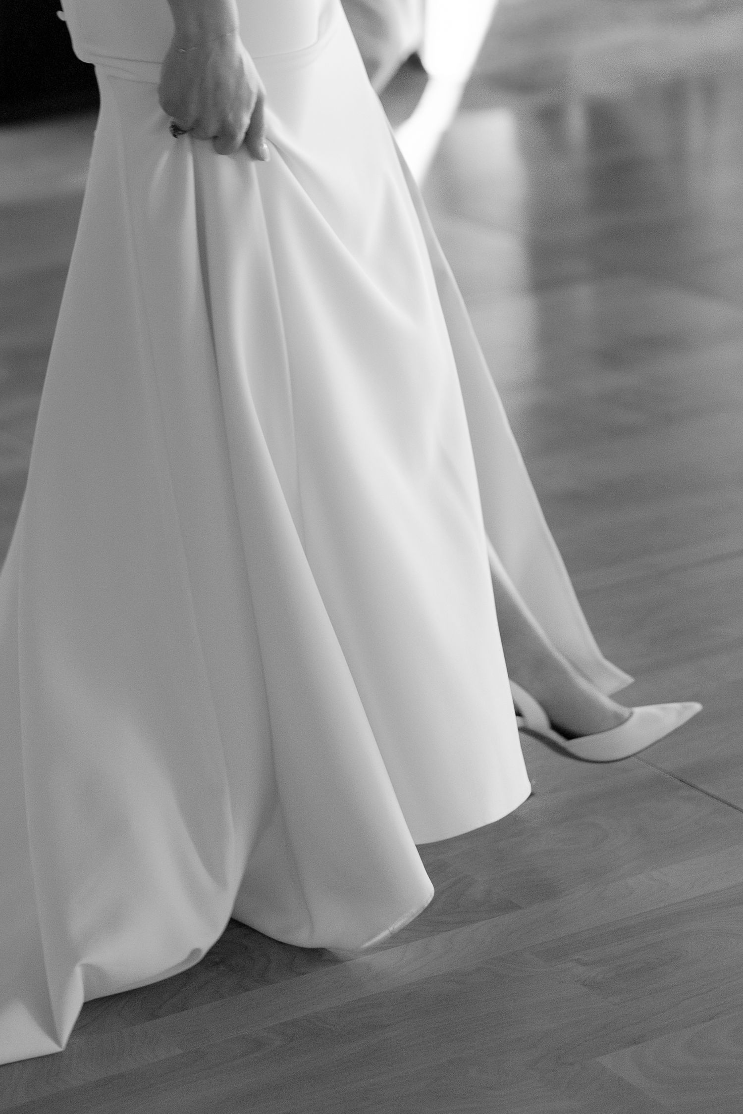 Close-up details of a flowing white wedding dress hemline against a dark floor.