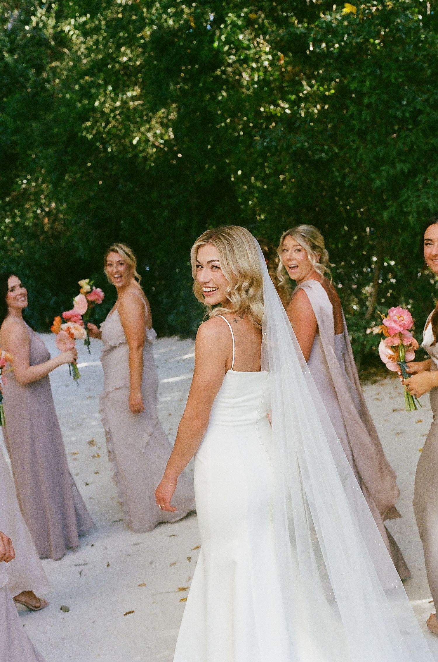 Wedding party in lavender bridesmaid dresses and white bridal gown pose outdoors on a sunny day.