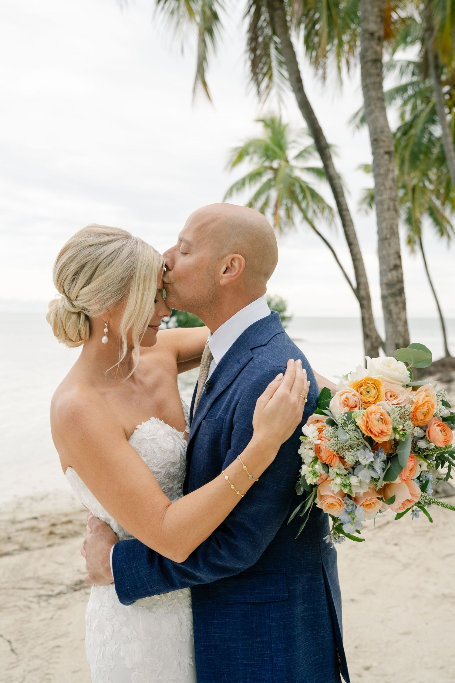 A romantic beach wedding moment with palm trees and peach-colored bouquet.