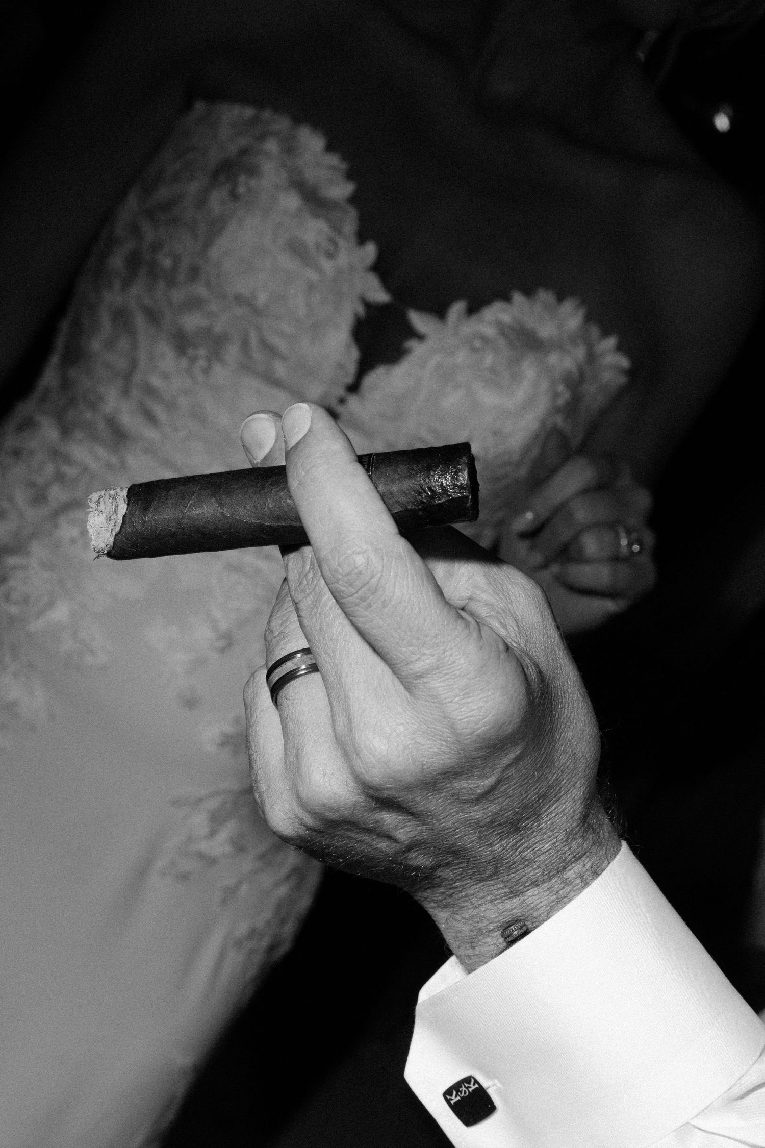 Black and white photo of hand holding cigar with wedding ring detail against lace dress.