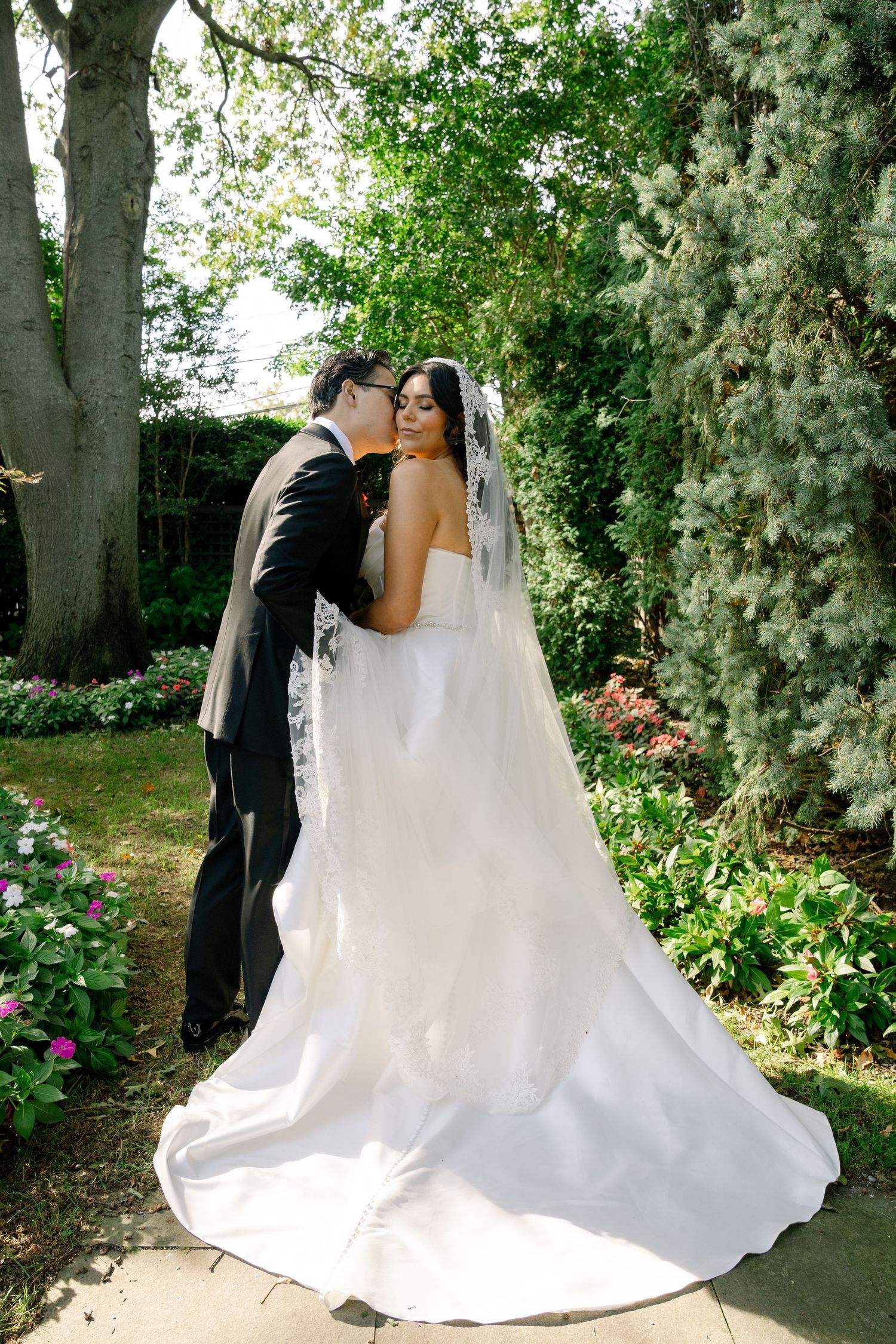 A romantic wedding couple embraces on a garden path surrounded by trees and flowering bushes.