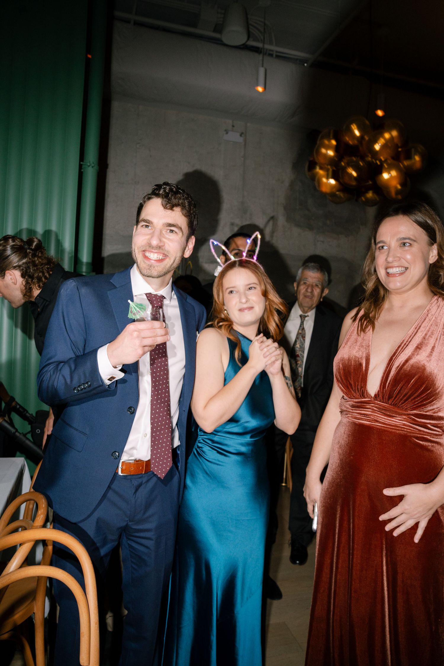 Party guests in formal attire enjoying themselves at an event with green curtains and ambient lighting.