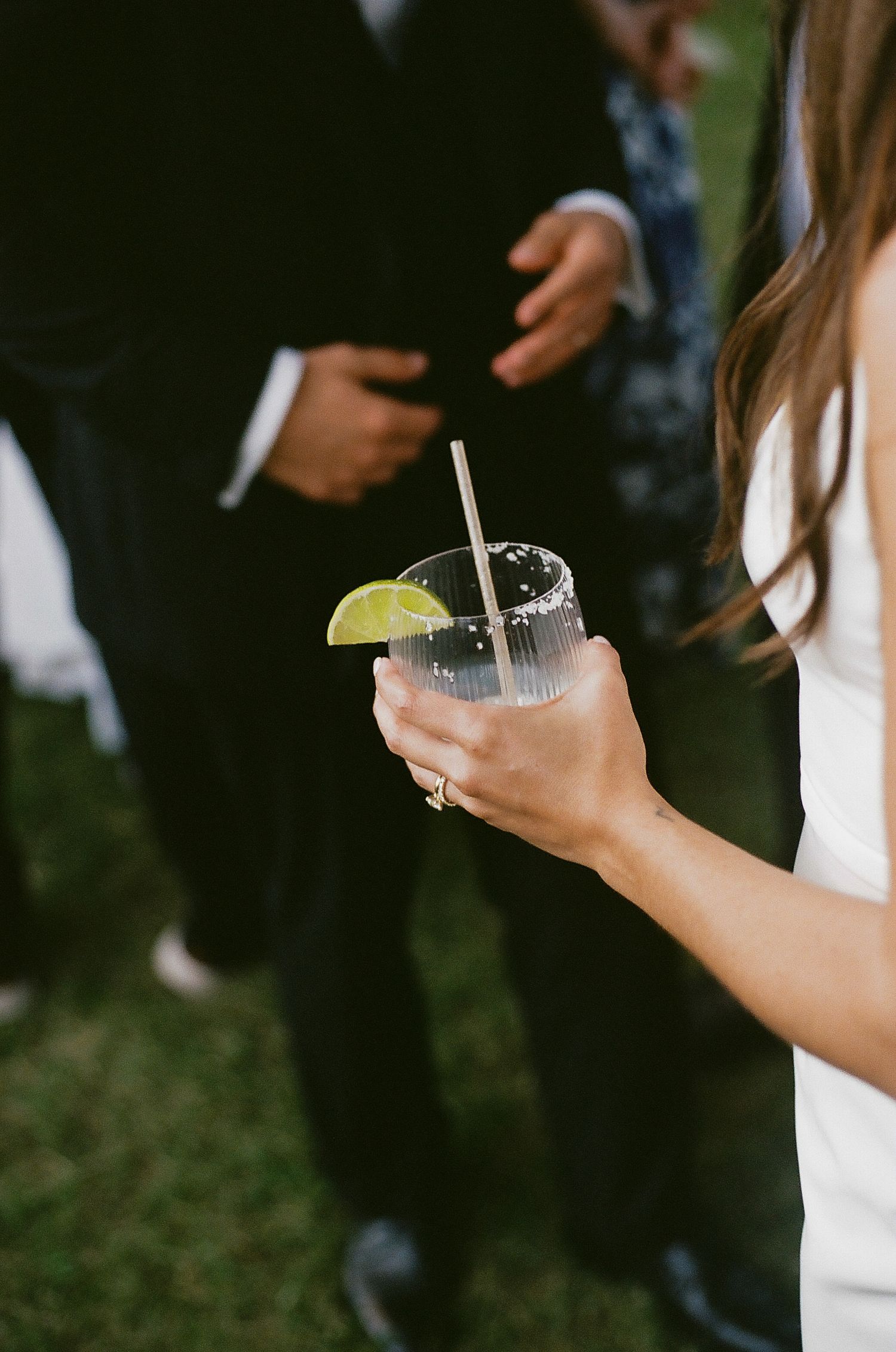 Bride holds a refreshing cocktail with lime garnish and straw during outdoor wedding celebration.