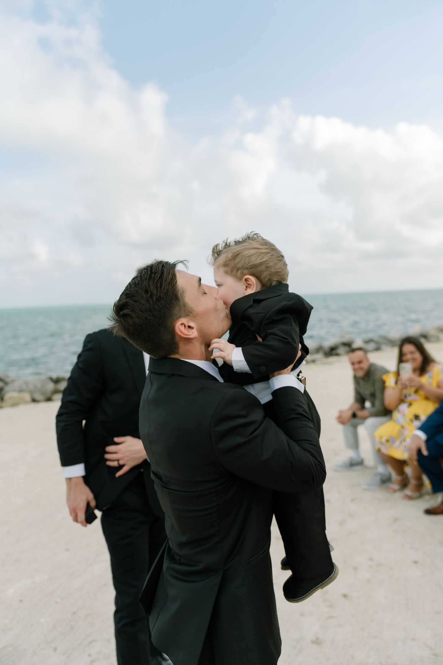 Man in black suit kissing a young boy in matching suit at a beachside wedding ceremony.