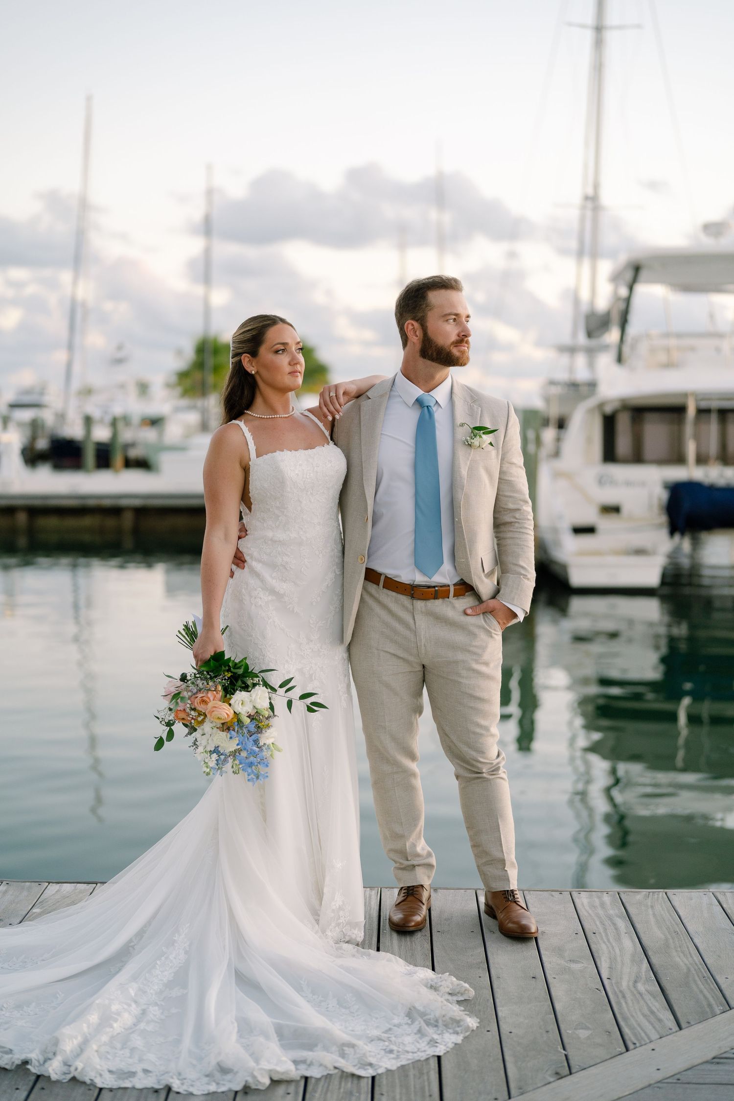 A wedding couple poses on a wooden dock with boats and marina views in the background.