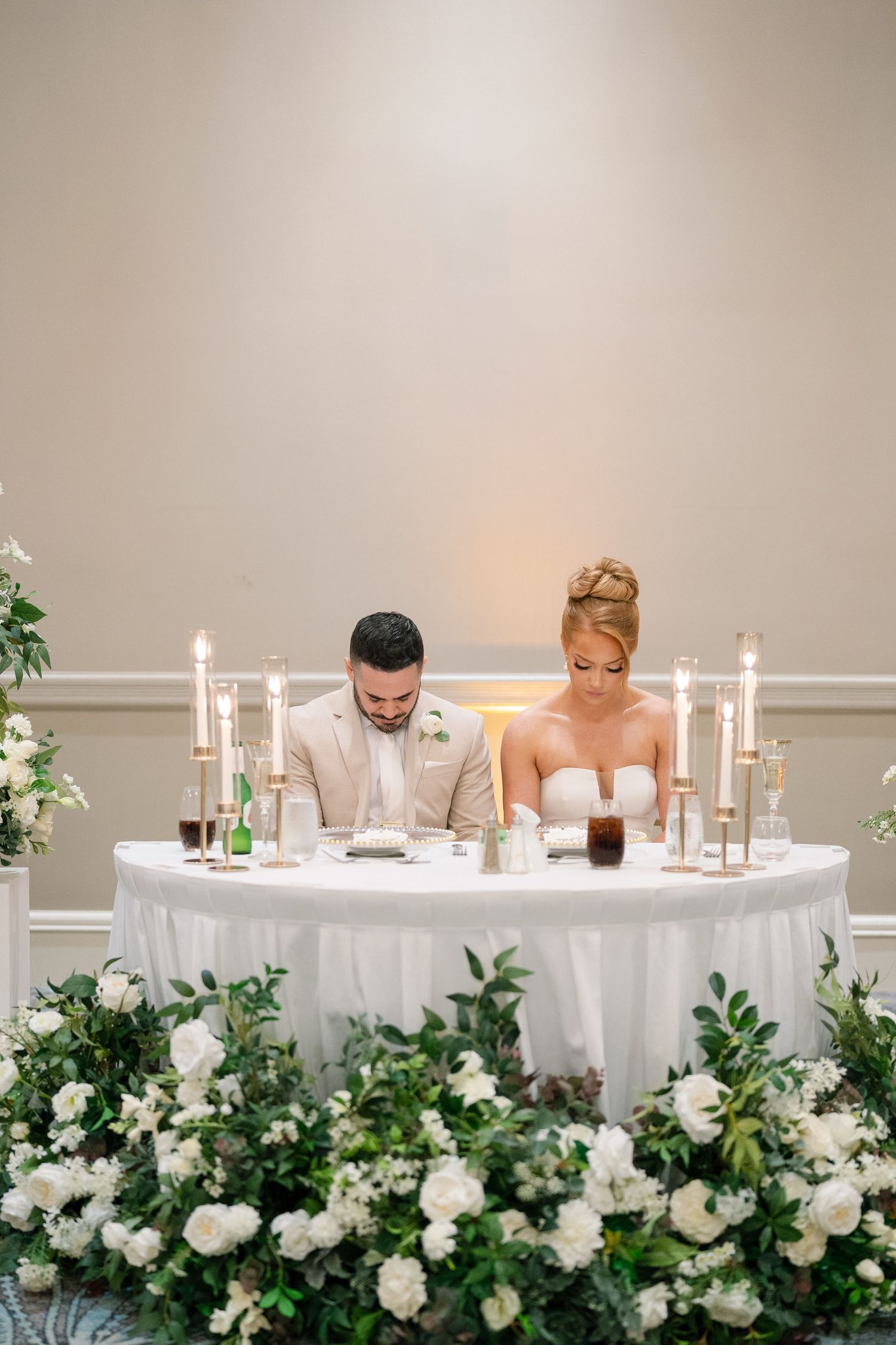 Wedding reception head table decorated with white flowers and candles against neutral wall.