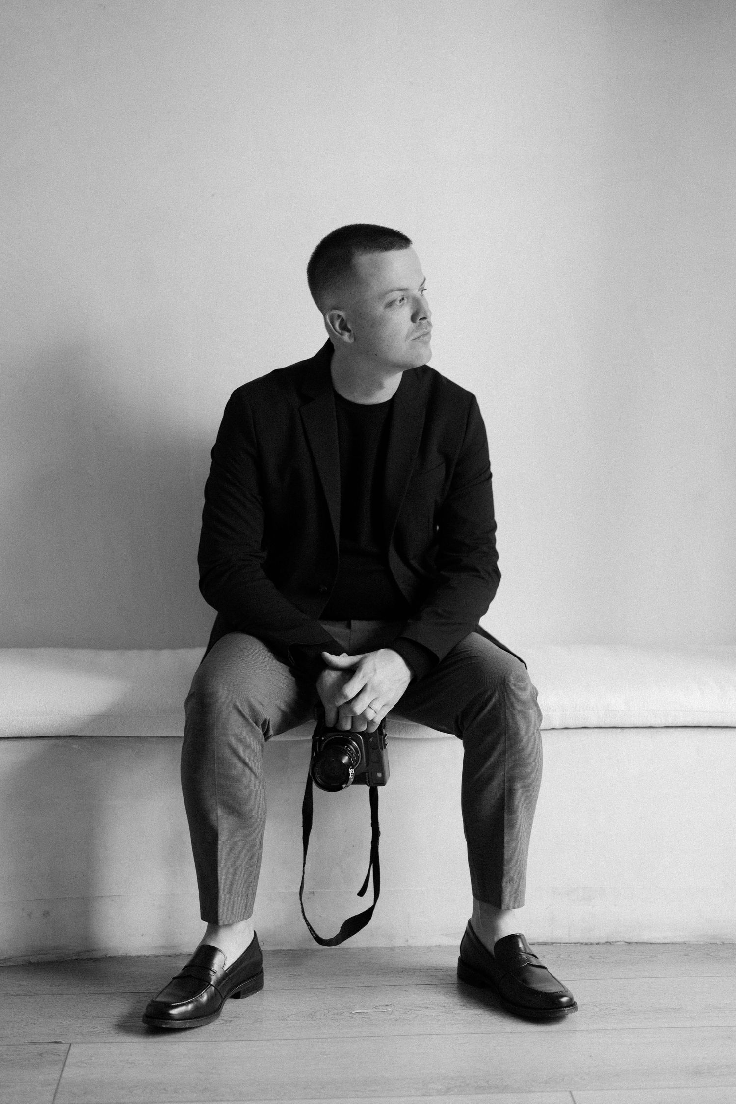 Person in black suit sits thoughtfully on floor against white backdrop in artistic portrait.