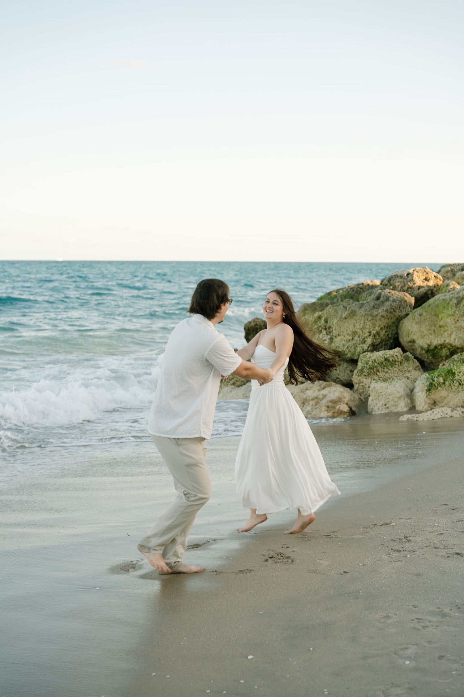 A couple dances playfully together on a beach with ocean waves and rocky shoreline in the background.