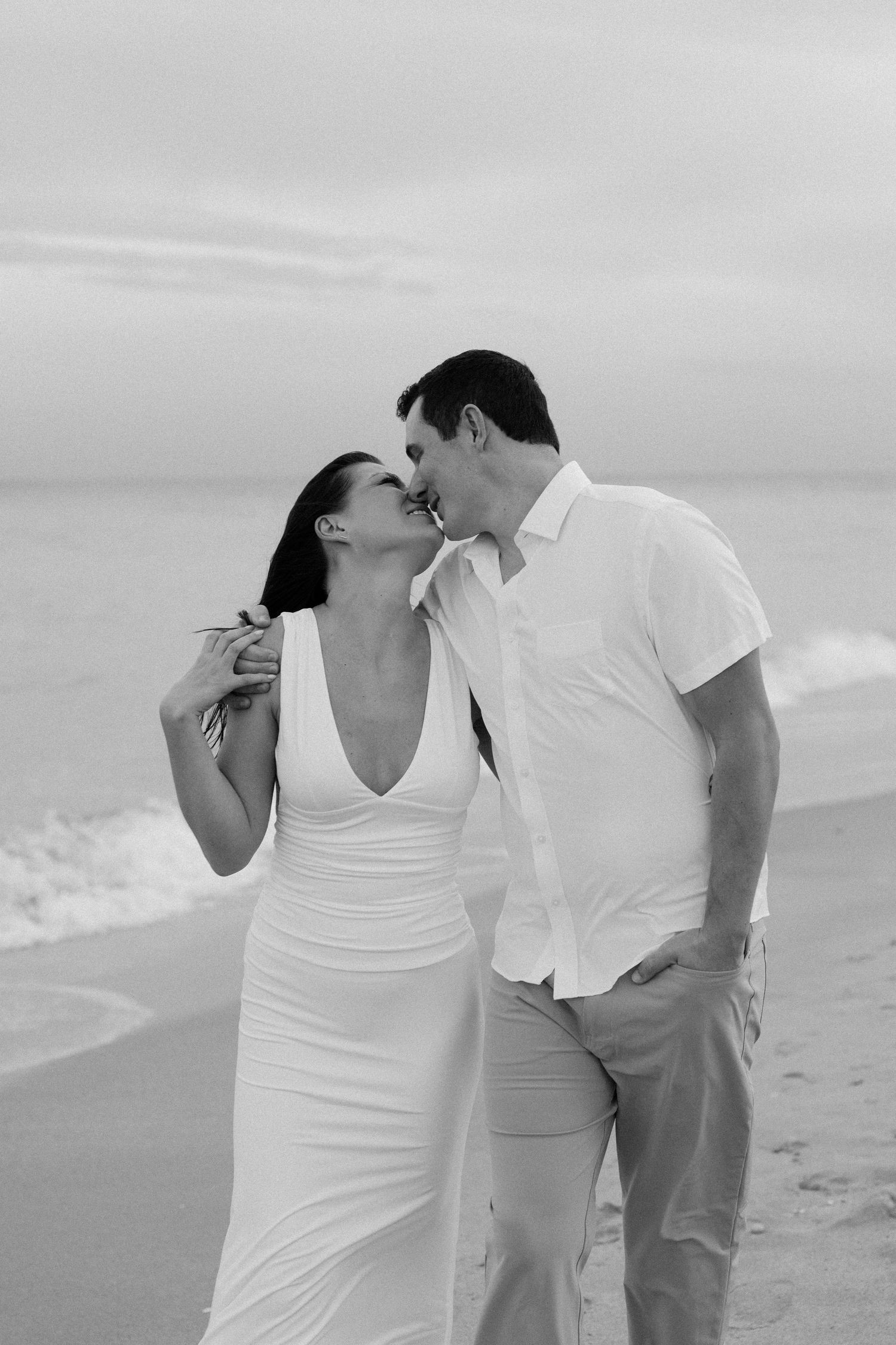 A romantic black and white beach photoshoot showing tender moments between a couple in white attire.