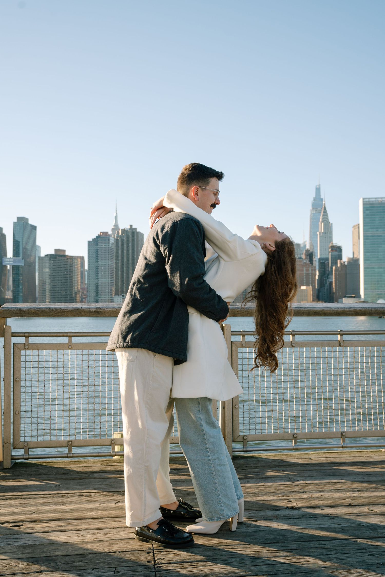 A couple dances romantically along the waterfront with the New York City skyline in the background.