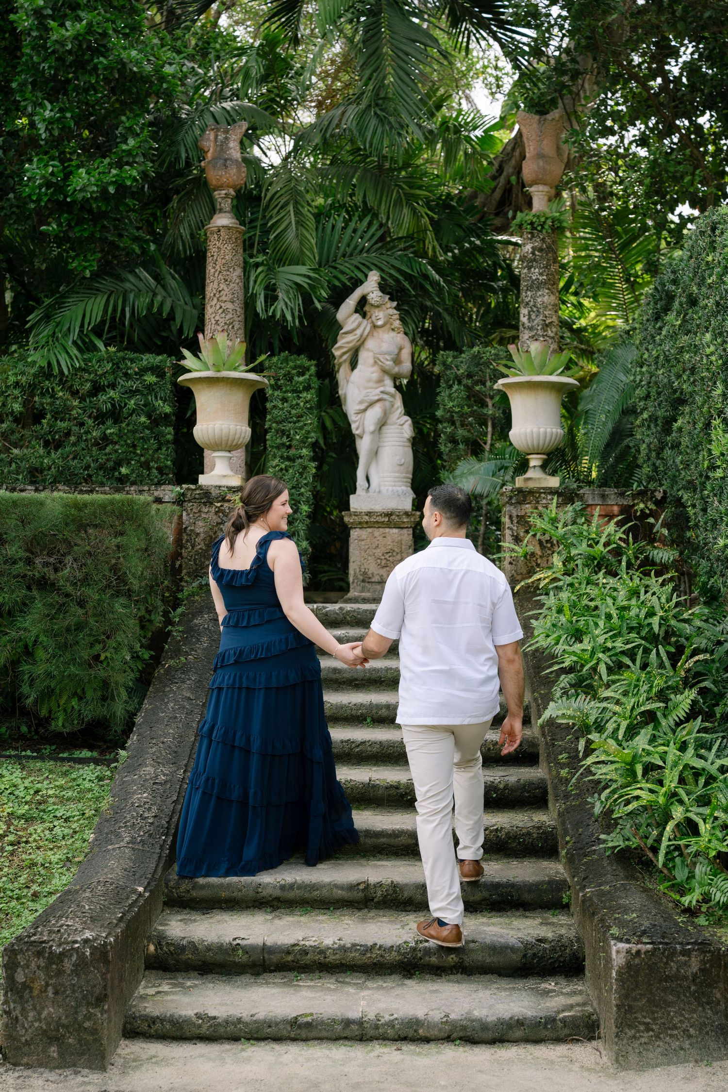 A couple ascends stone steps flanked by classical statues and ornate pillars in a tropical garden.