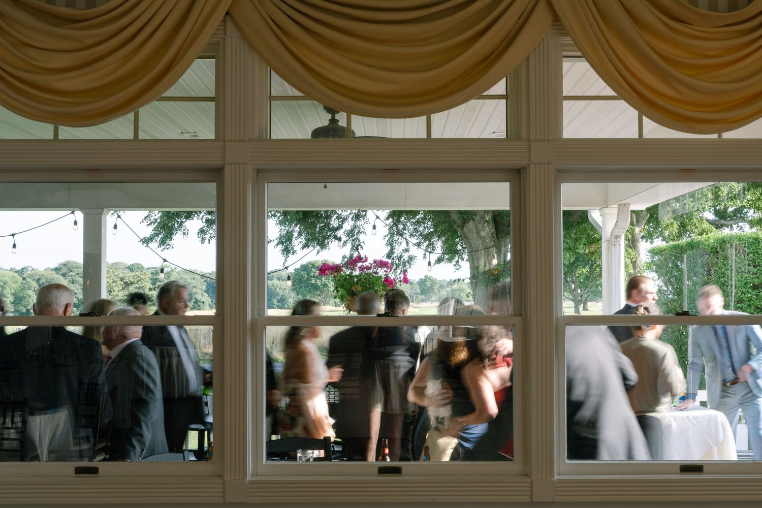 Guests mingle inside an elegant banquet hall viewed through large windows with gold draping.