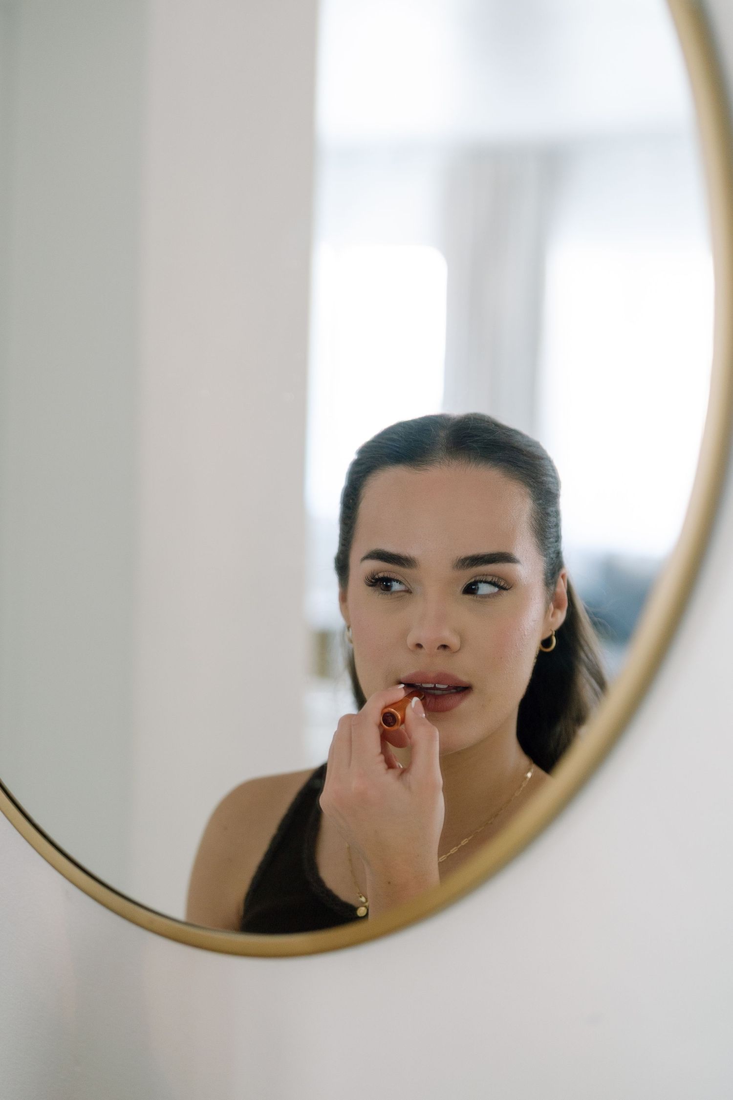 Young woman applying red lipstick while looking at her reflection in a round gold-framed mirror.