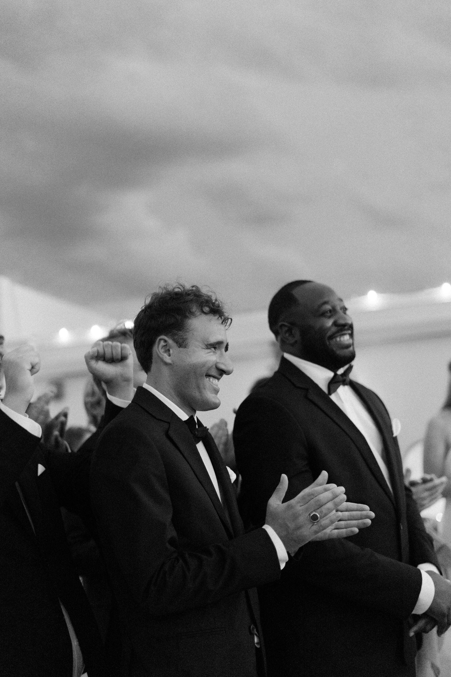 Wedding party members in formal black attire share laughs during an outdoor celebration.