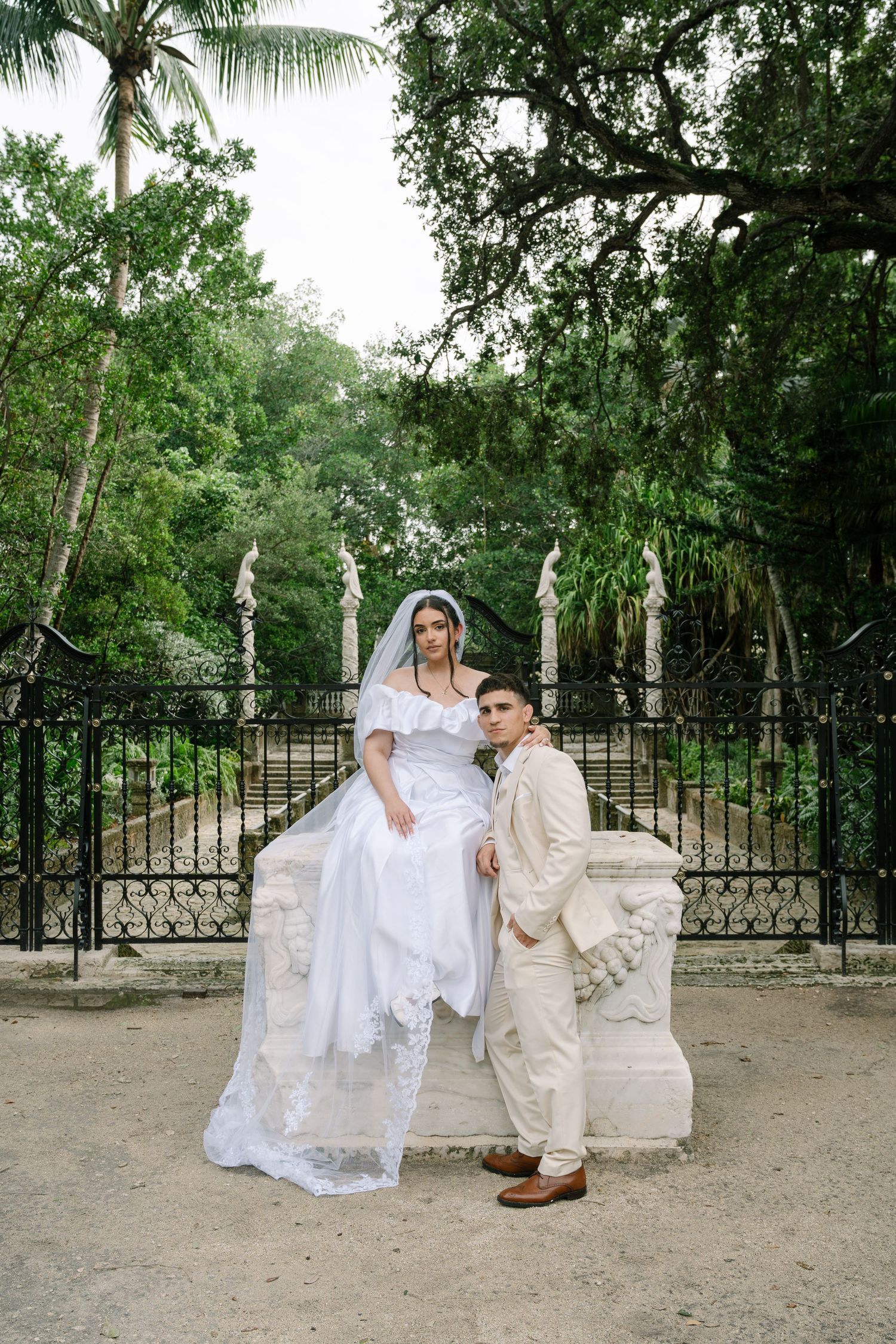 A couple poses in wedding attire in front of an ornate iron gate surrounded by lush tropical foliage and palm trees.