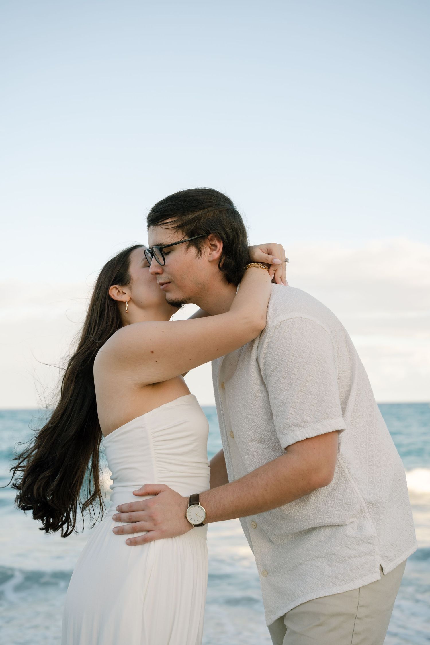 A couple embraces tenderly during a beach photoshoot with the ocean and soft sky behind them.