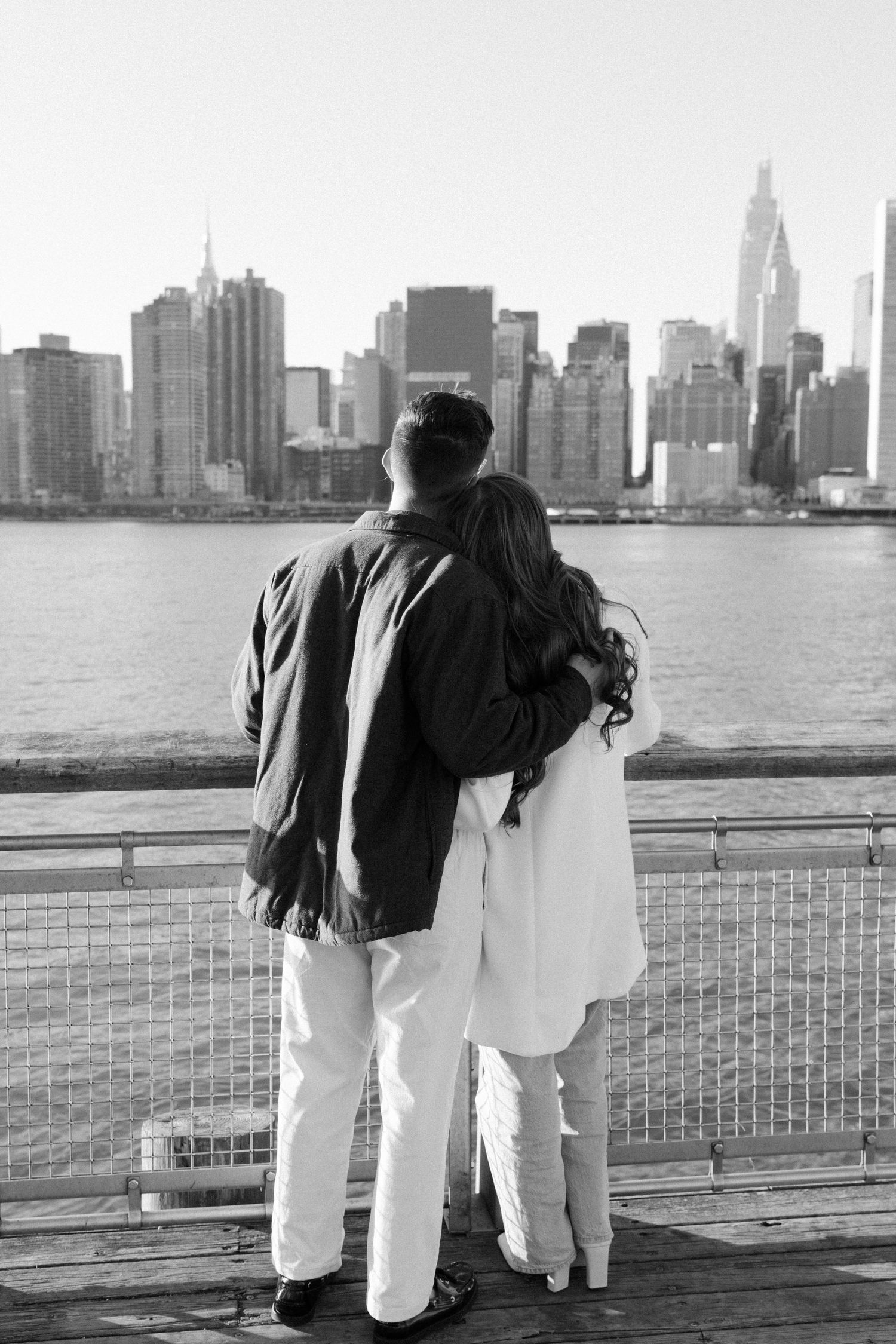 A couple stands at a railing overlooking the New York City skyline across the water in black and white.