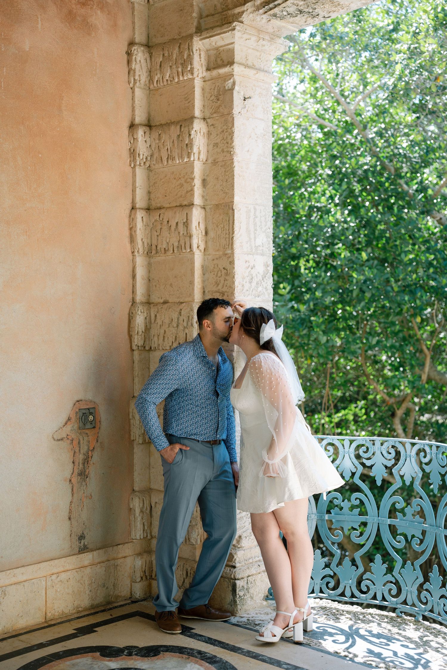 A couple shares a romantic moment in an ornate stone colonnade with lush greenery visible in the background.