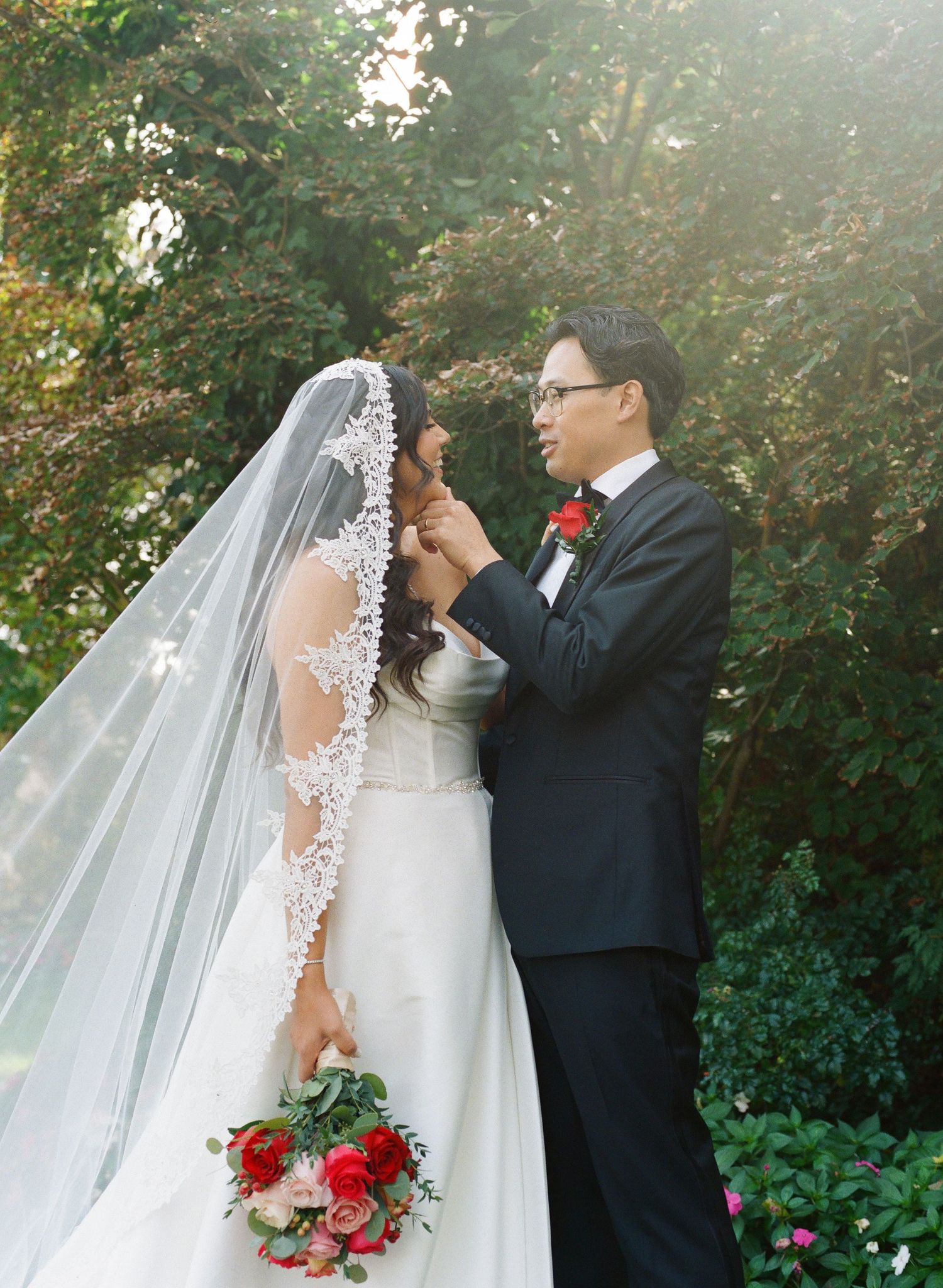 A romantic wedding couple shares an intimate moment in a garden setting with lush greenery and sunlight filtering through.