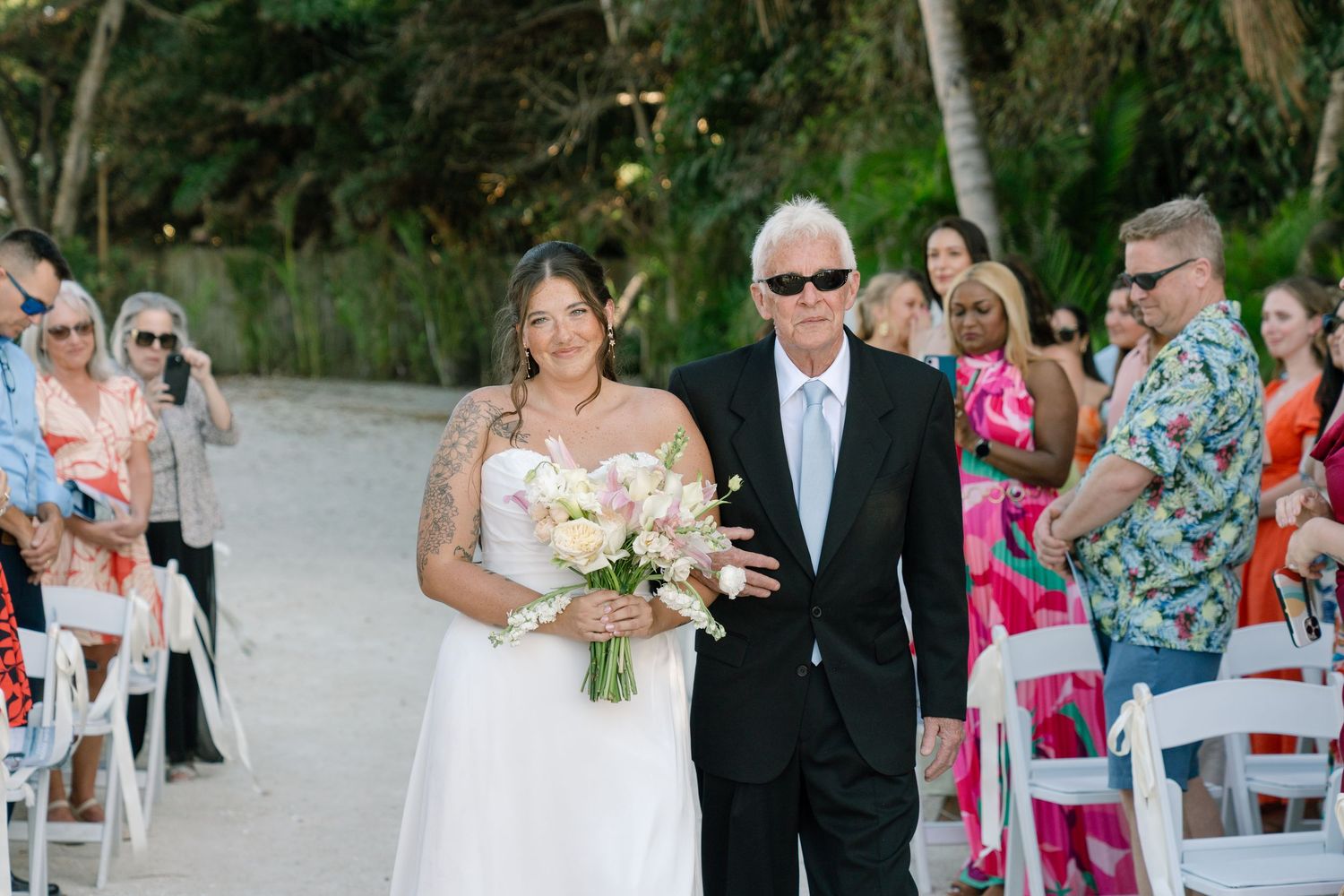 Bride in white gown walks down sandy beach aisle with escort holding pink bouquet at outdoor wedding ceremony.