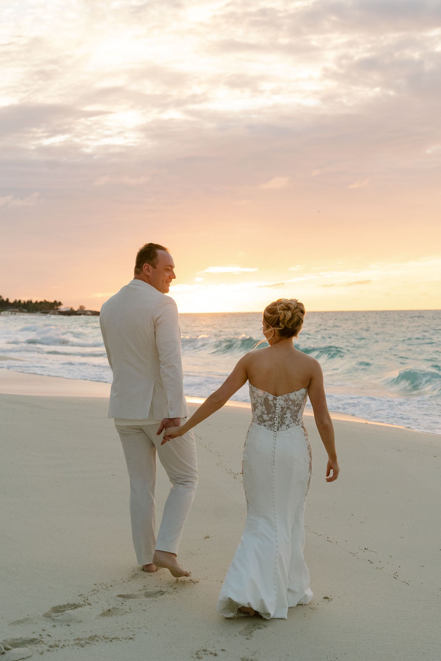 Bride and groom holding hands walking on a sandy beach at sunset, bride in a fitted lace gown.
