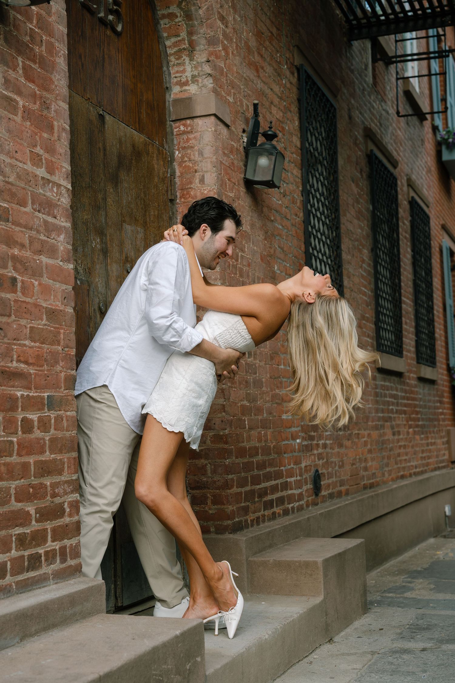 Couple dancing romantically on brick building steps, man in white shirt dipping blonde woman in white dress and heels.