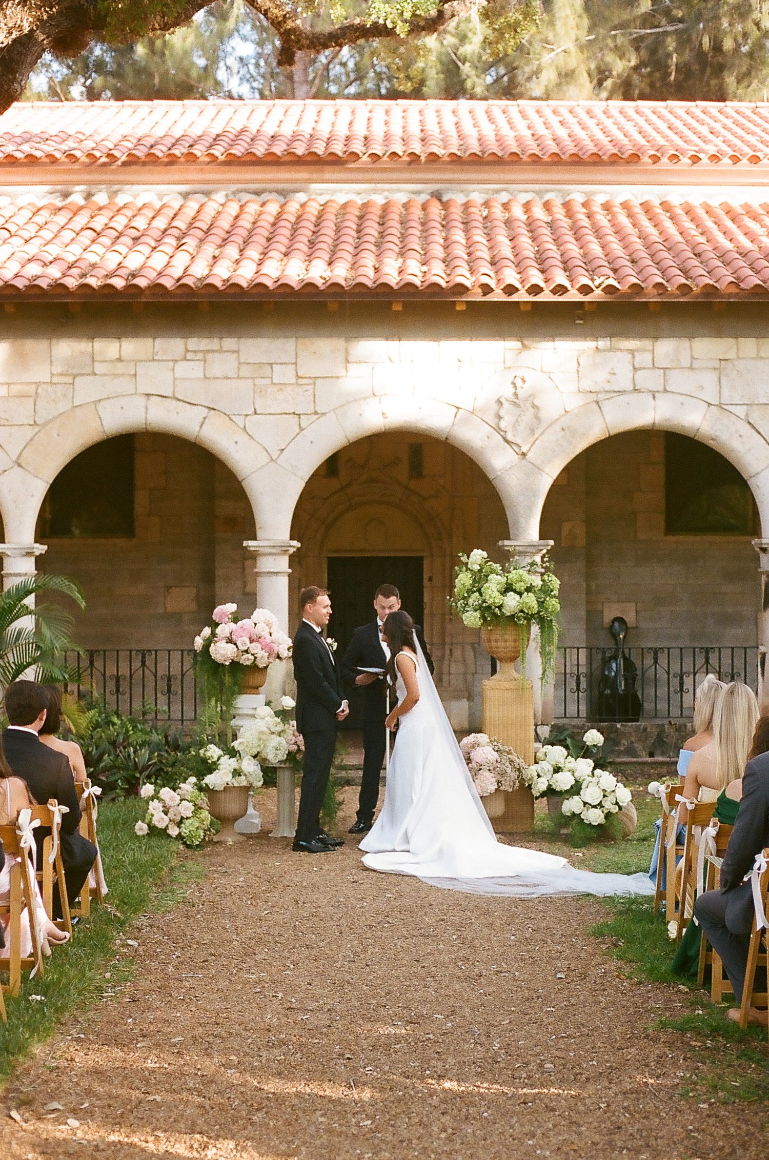 Outdoor wedding ceremony at a Spanish-style stone chapel with floral arrangements and seated guests.